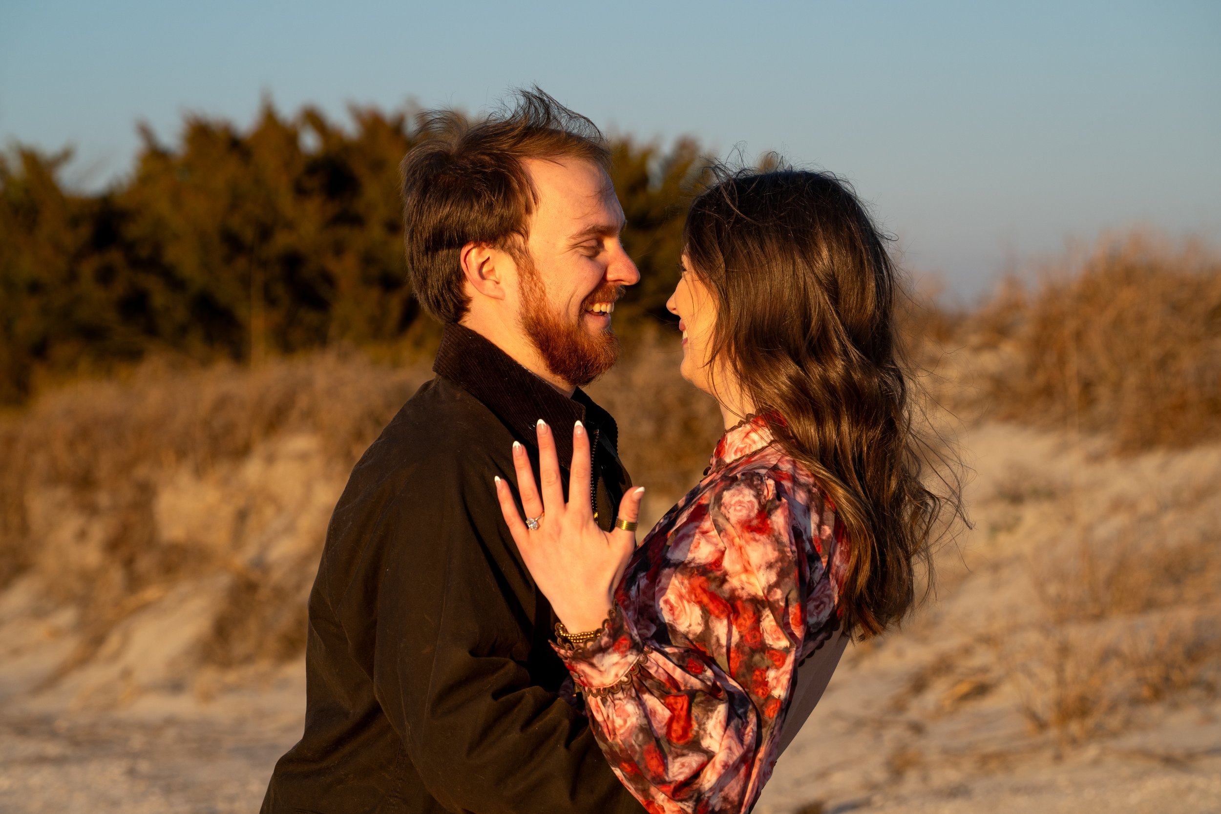 A couple smiling and facing each other on a beach at sunset, with desert shrubs and blue sky in the background.