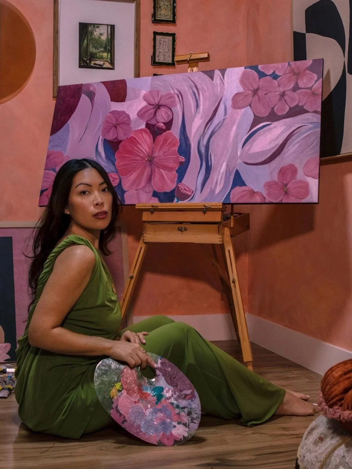 A woman with long dark hair sitting on a wooden floor holding a colorful paint palette, in front of a pink wall with framed artwork and a large canvas of pink flowers on an easel.