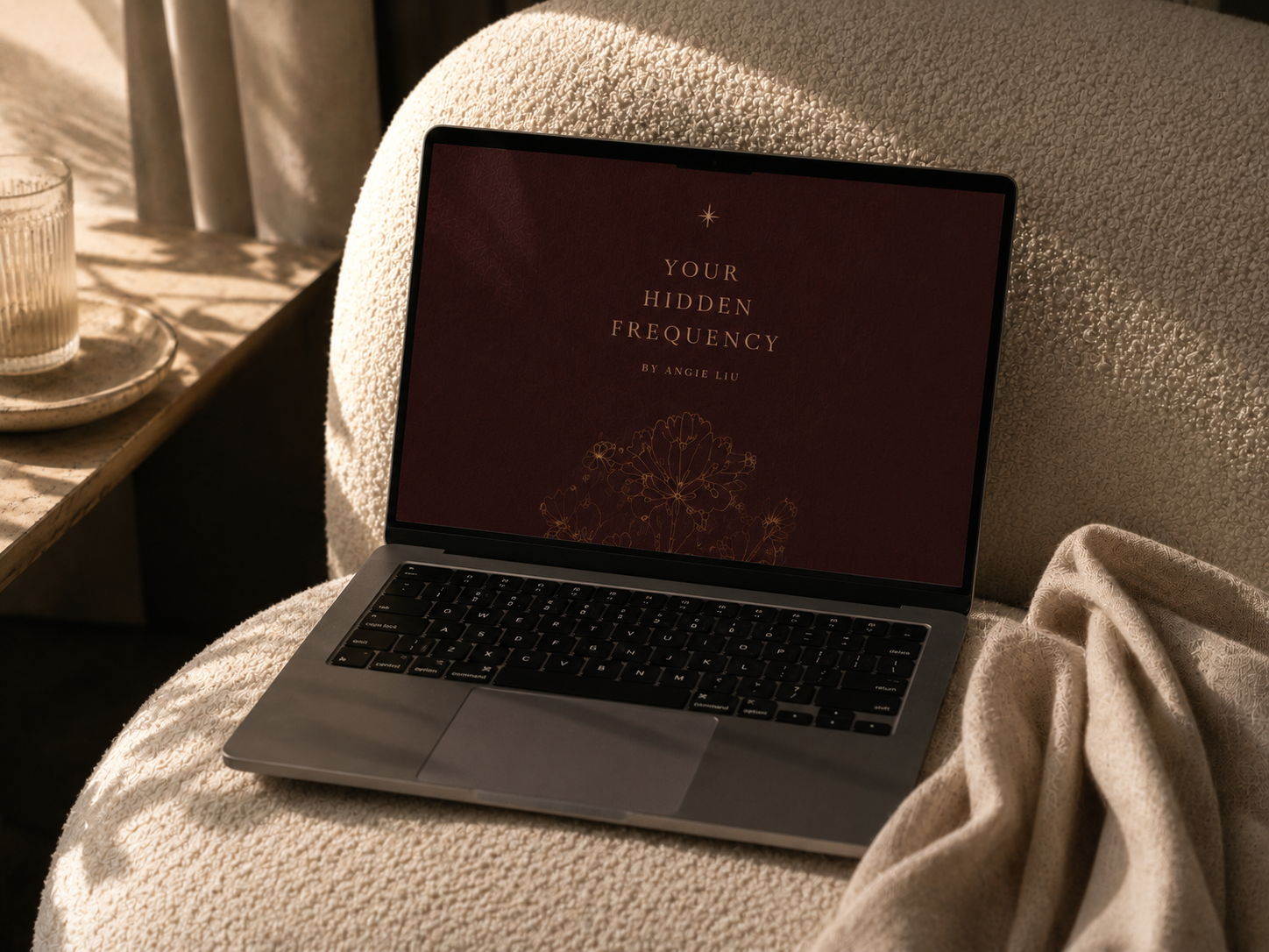 A laptop on a cozy, textured beige blanket reads 'Your Hidden Frequency' by Angie Liu with a floral design on the screen.