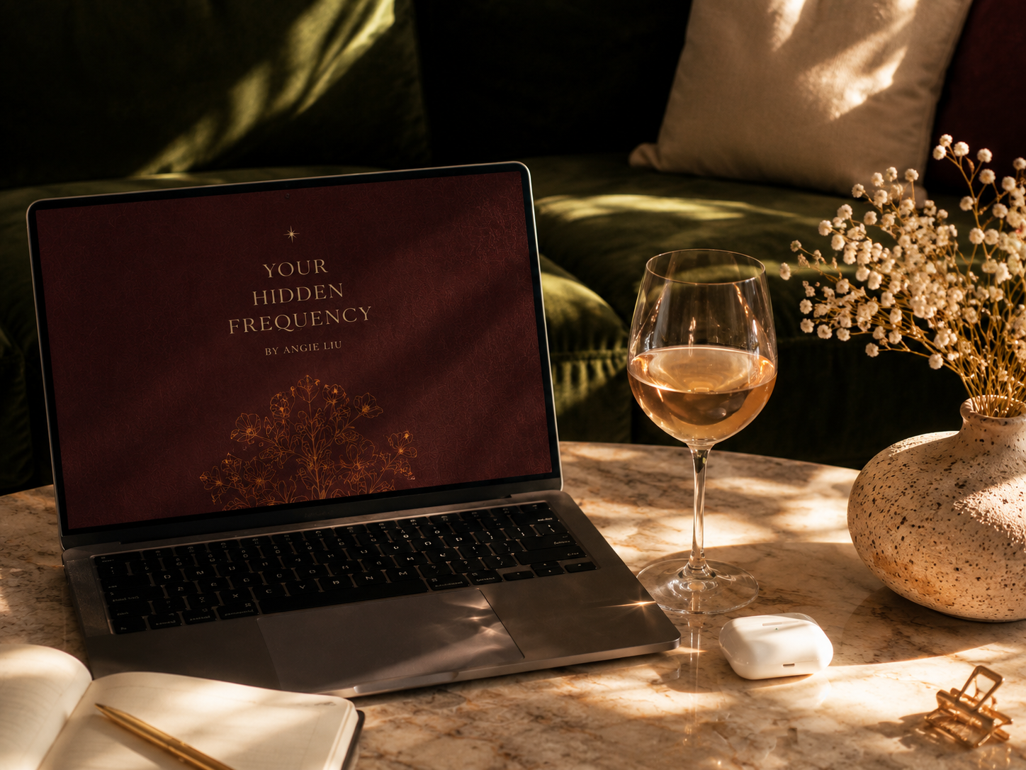 A workspace setup on a marble table featuring a laptop displaying the title 'Your Hidden Frequency' by Angie Liu, a glass of white wine, a white vase with white baby's breath flowers, an open notebook with a gold pen, and a small white wireless earbud case. In the background, there is a dark green velvet sofa with pillows.
