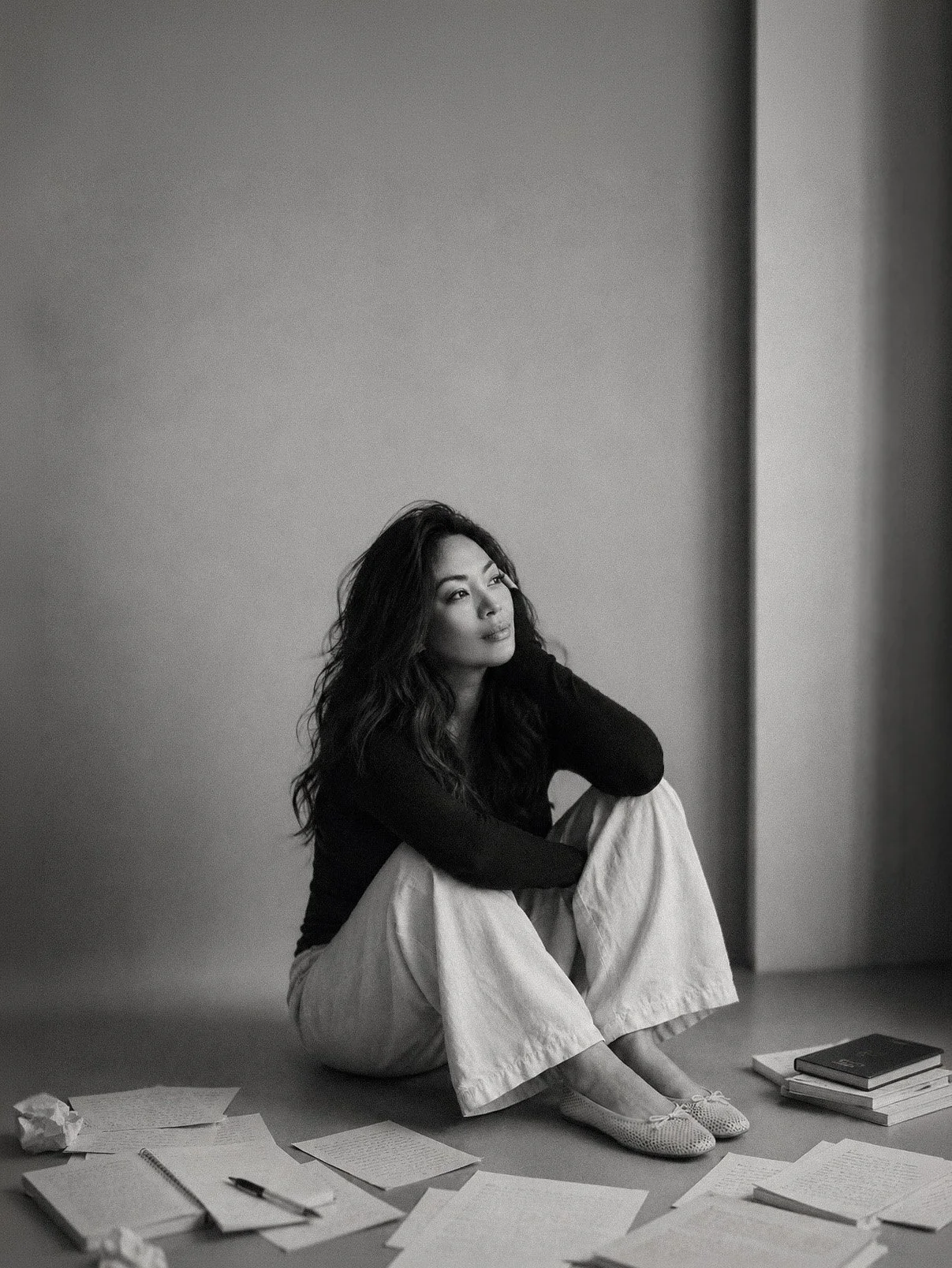 A black-and-white photo of a woman sitting on the floor, surrounded by scattered papers, books, and notebooks. She has long wavy hair and is wearing a dark top and light-colored pants, looking contemplative.