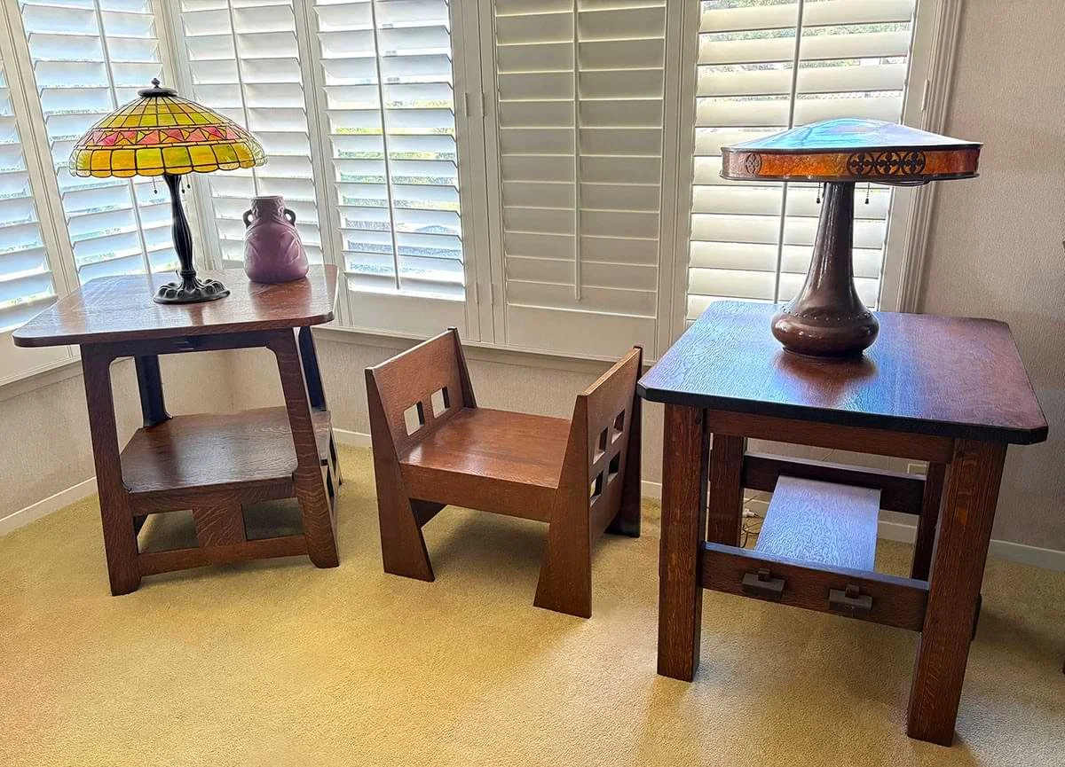 Two wooden tables with lamps and a small wooden chair in front of a window with white shutters.