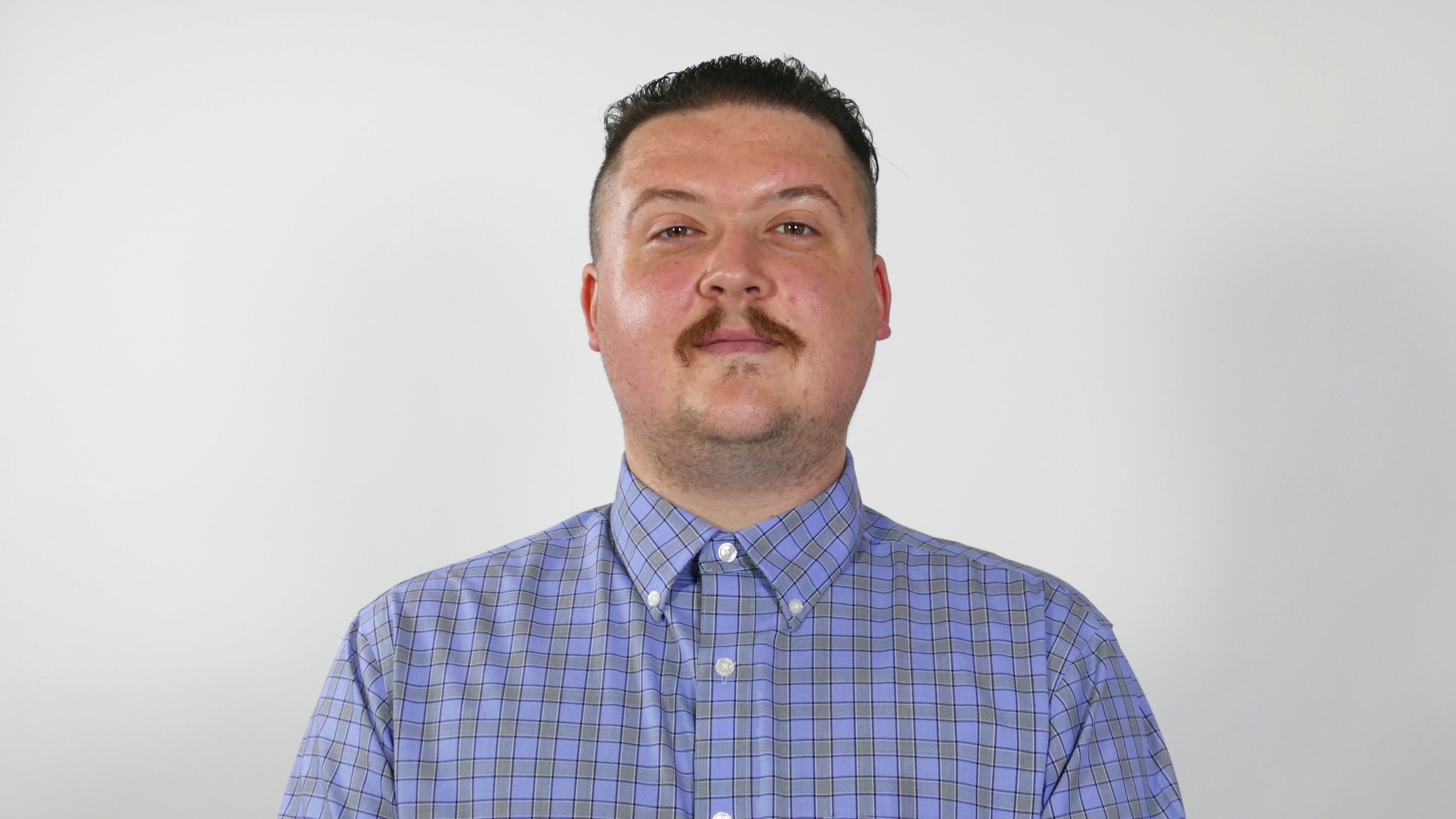 Portrait of a man with short dark hair, facial hair, wearing a blue checkered button-up shirt, against a plain white background.