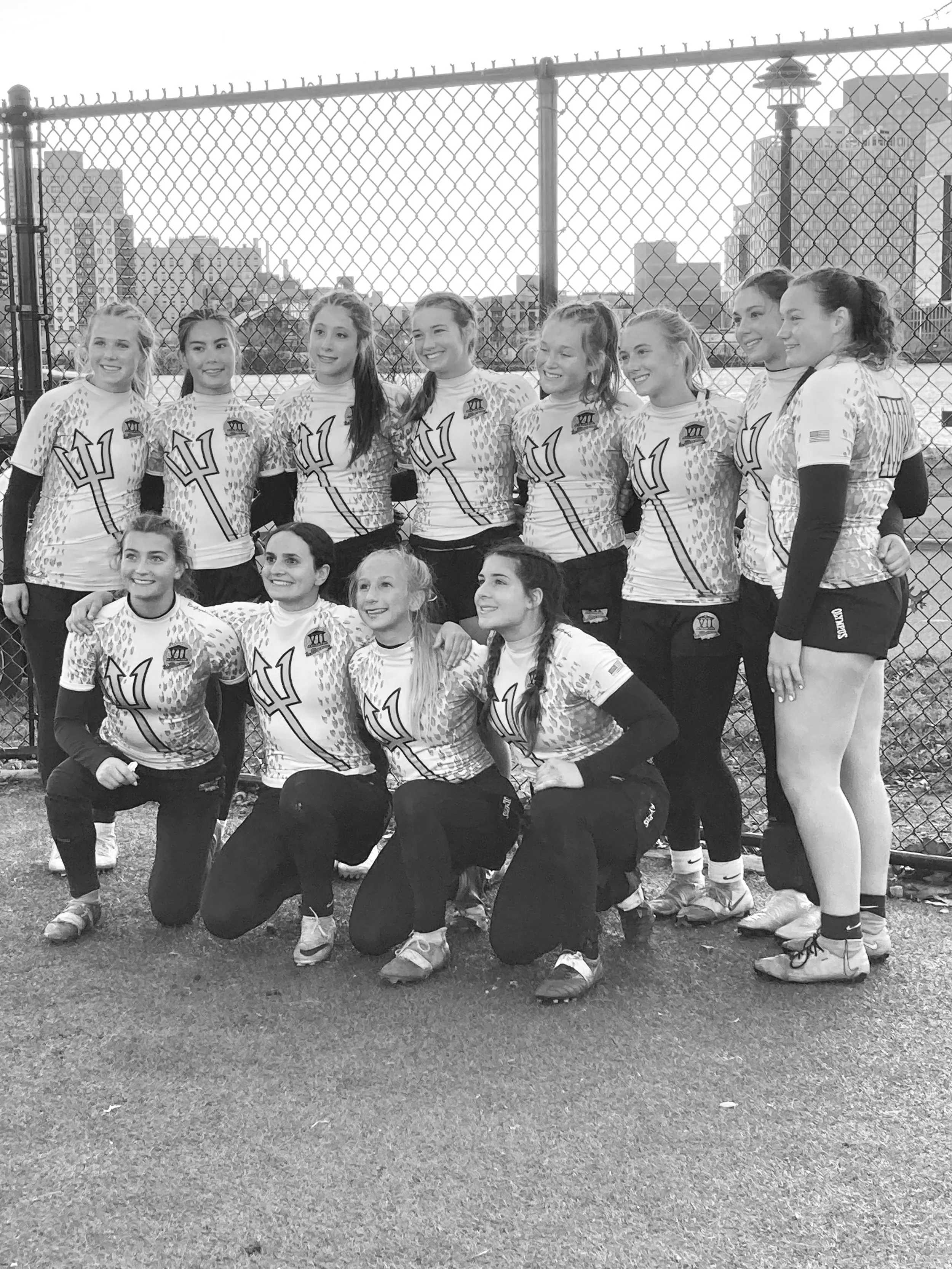 A group of young women in athletic uniforms posing outdoors in front of a chain-link fence with city buildings in the background. They are smiling and appear to be on a sports team.
