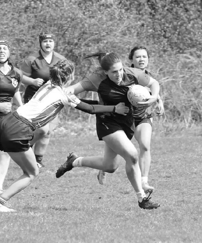 Women playing rugby outdoors, with one player holding the ball and others reaching for her.