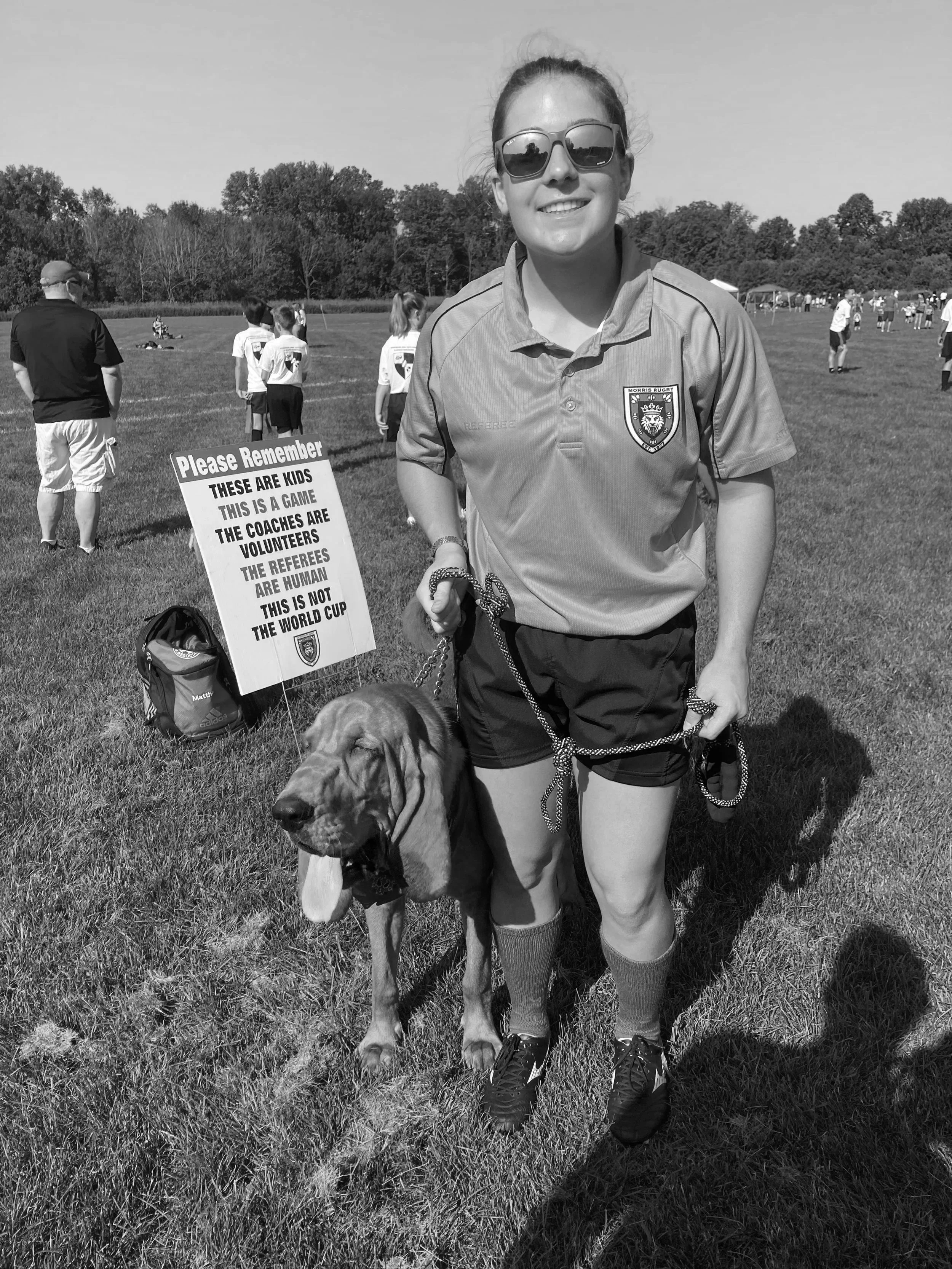 sunglasses, wearing a sports uniform, stands on a grassy field holding a leash attached to a large dog. In the background, there are people and a sign that reads: "Please Remember These Are Kids. This Is a Game. The Coaches Are Volunteers. The Referees Are Human. This Is Not the World Cup."