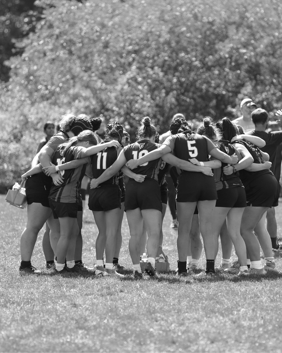 A group of female rugby players huddled together with their arms around each other on a field, with trees in the background.