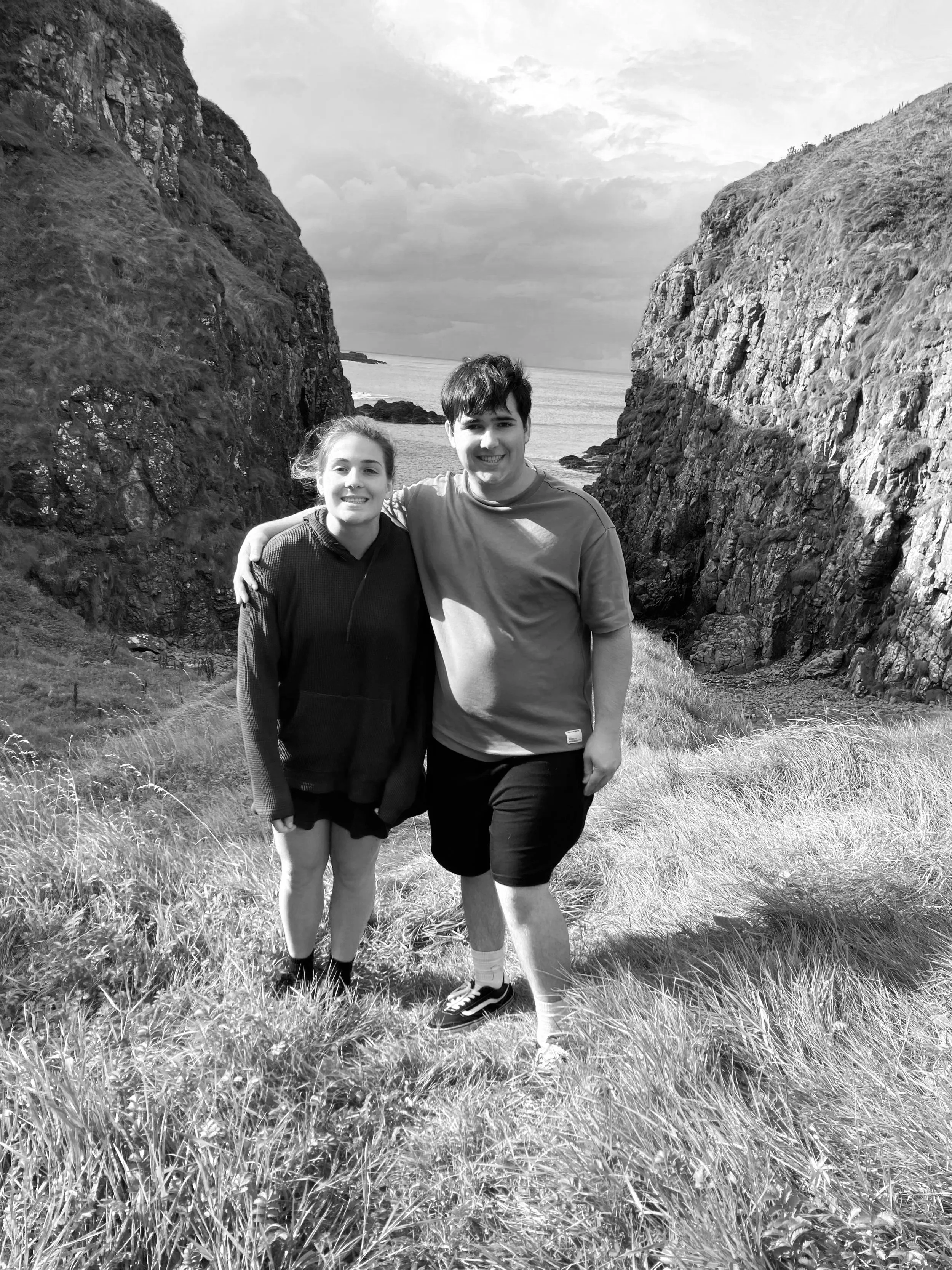 Two young people, a girl and a boy, standing outdoors between large rock formations with grass on the ground, smiling at the camera. In the background, there are cliffs and a body of water under a cloudy sky.