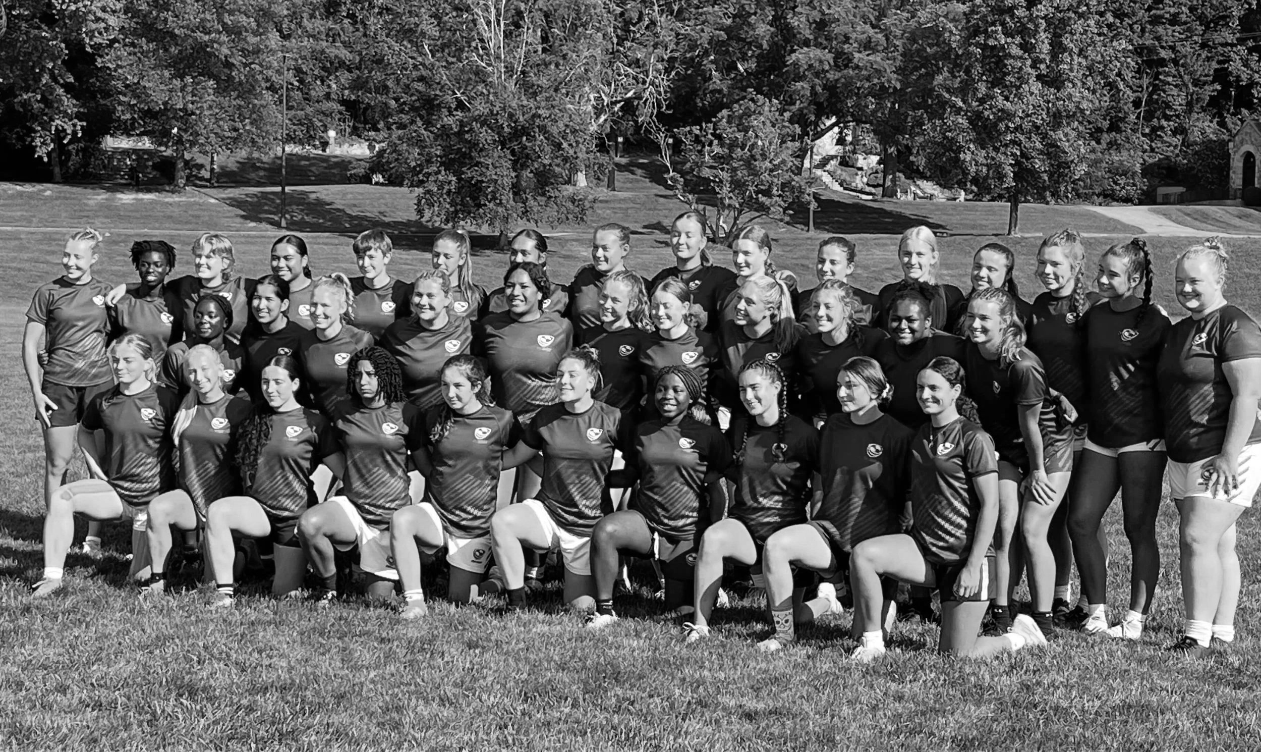 A black and white photo of a women's sports team posing outdoors on grass, with trees and buildings in the background. The team members are in uniform, standing and kneeling in two rows.