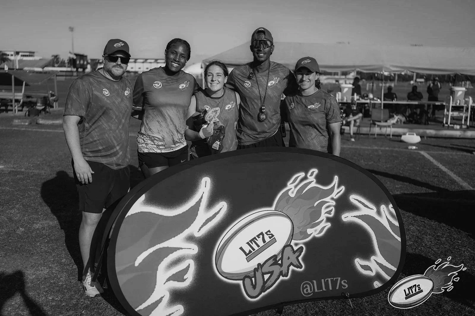 Group of five people standing behind a lit-up sign with a flaming football graphic that says 'LITZ USA.' They are at an outdoor sports event under a tent.