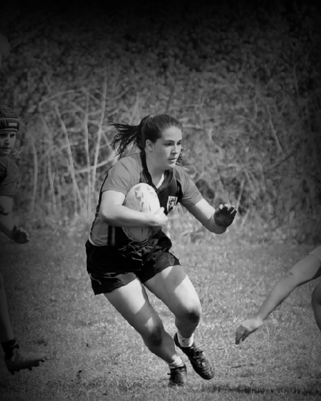 A woman playing rugby on a field, holding a rugby ball and running with determination, with two other players partially visible nearby.
