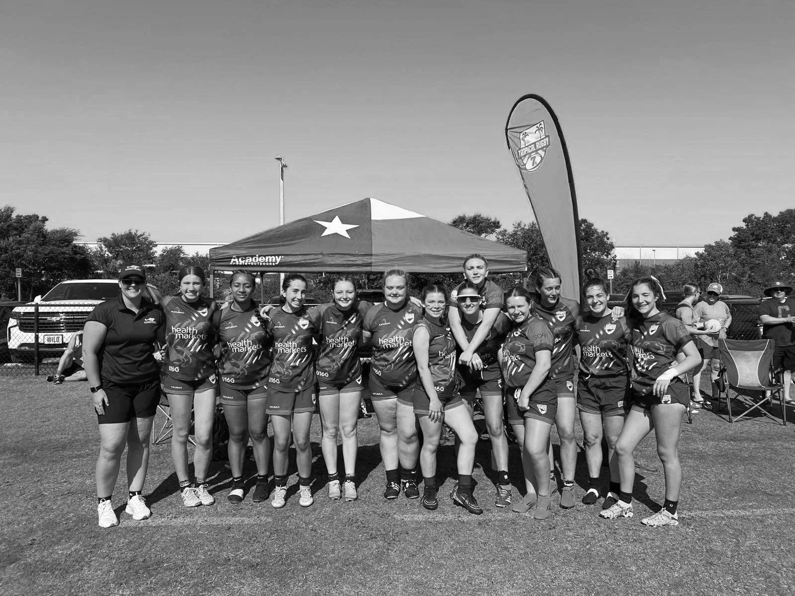 A group of young female athletes in sports uniforms standing outdoors with a coach, posing for a team photo at a sports event, under a tent with a flag and trees in the background, on a sunny day.