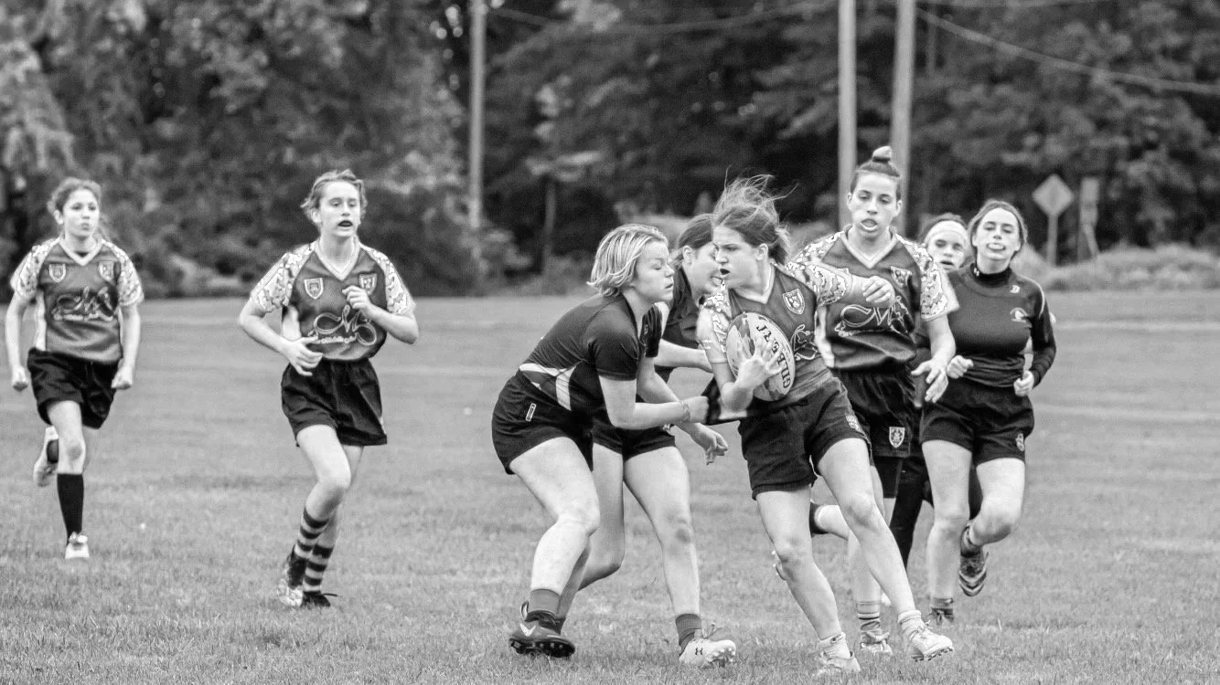 Black and white photo of girls playing rugby on a field, with one girl holding a rugby ball and others running around her.
