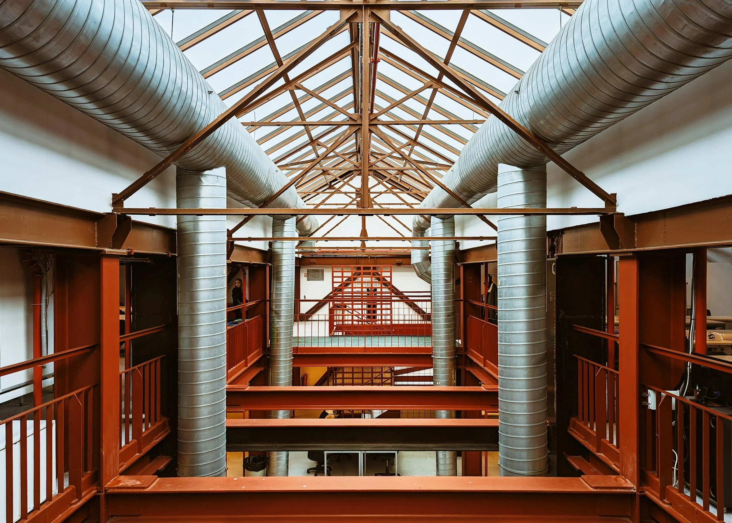 Interior view of a building with large metallic air ducts, red metal framework, balconies, and a glass ceiling above.