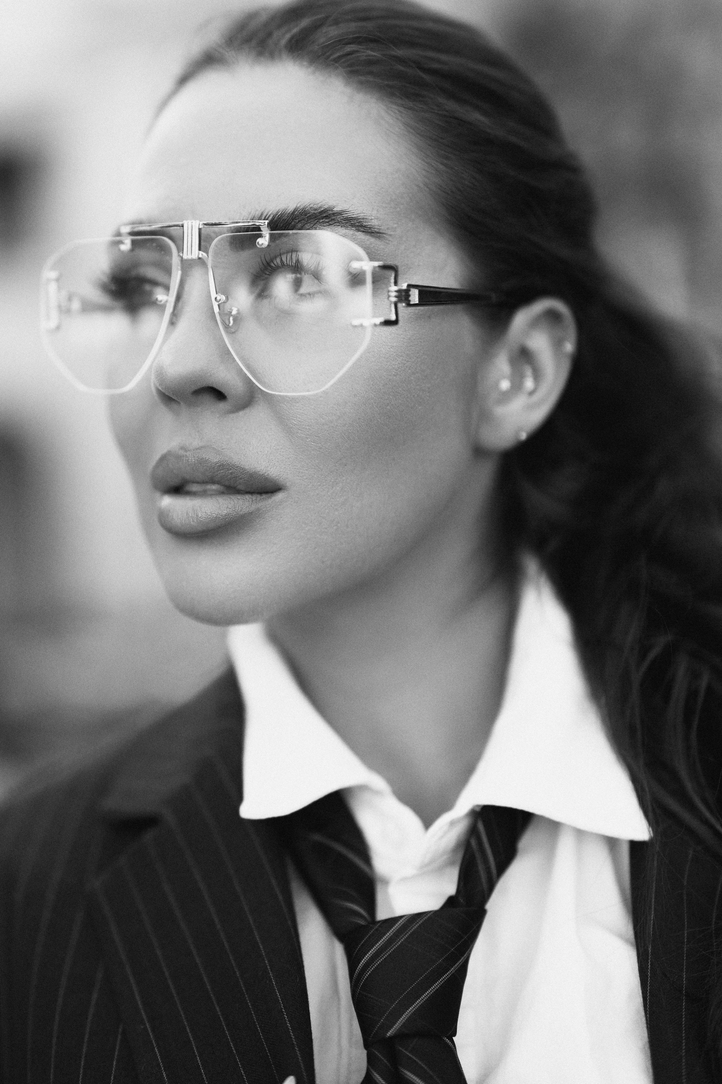 A woman with dark hair in a ponytail, wearing stylish large glasses, a pinstripe suit, white shirt, and tie, looking to her left in black and white.