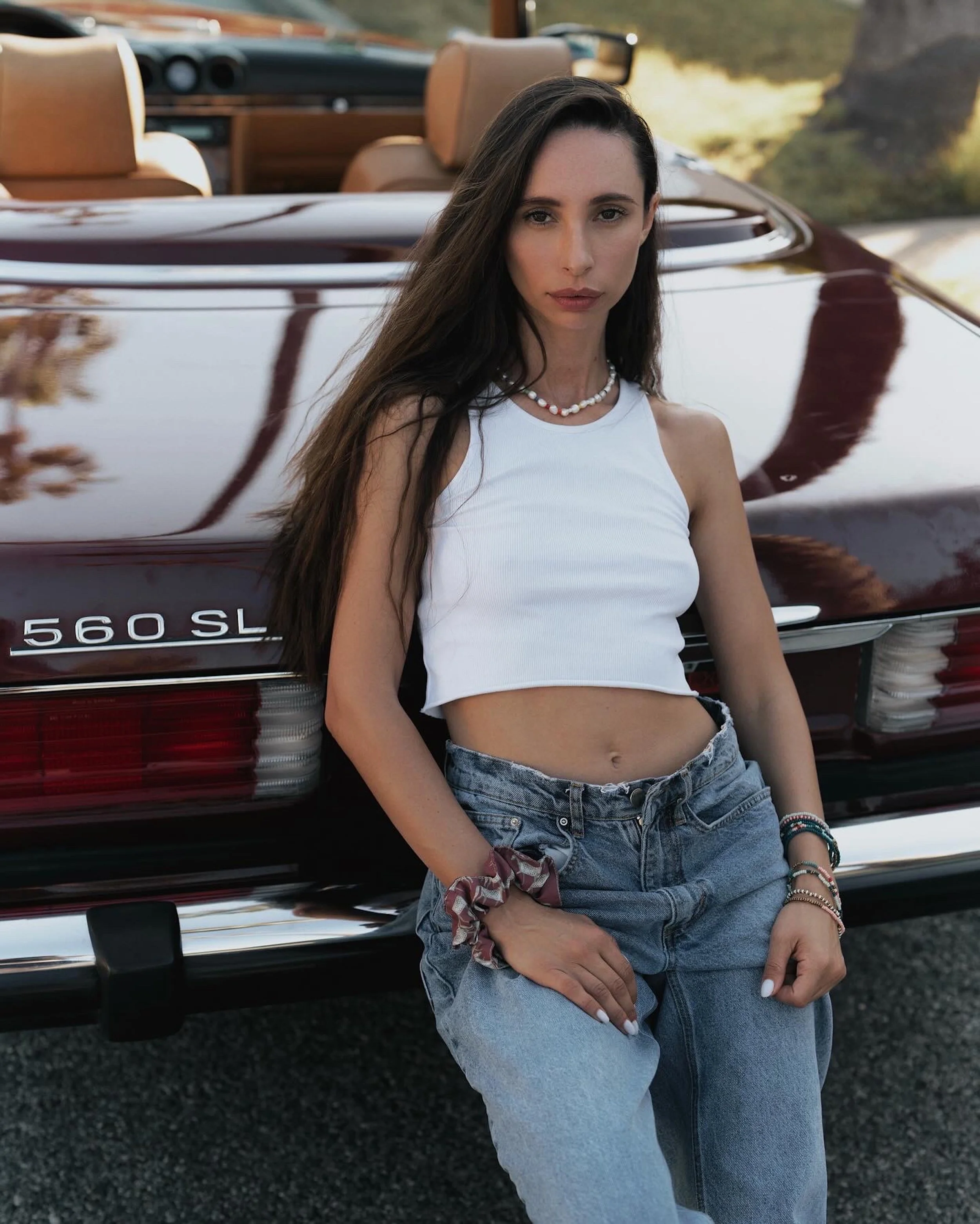 Young woman with long dark hair standing in front of a vintage Mercedes-Benz 560SL convertible car, wearing a white crop top and high-waisted jeans, with accessories on her wrist and neck, during daytime.