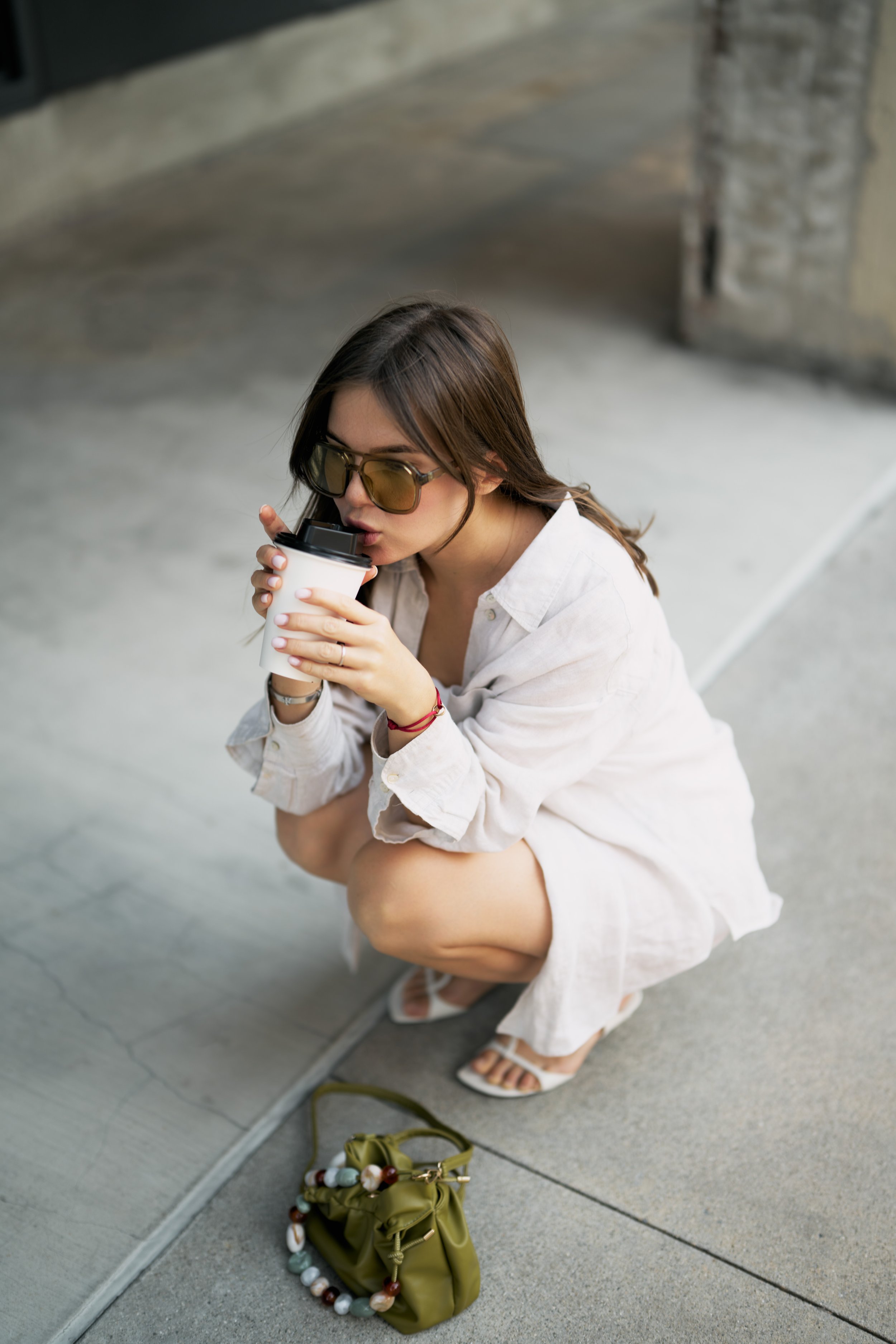 Woman squatting on sidewalk wearing white dress and sunglasses, drinking coffee from a to-go cup, with a green purse and necklace on the ground beside her.
