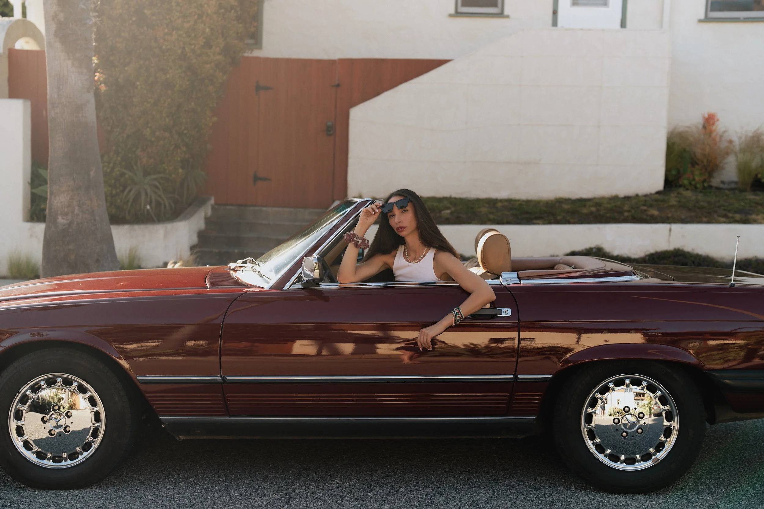 A woman with long dark hair, wearing sunglasses, a white tank top, and jewelry, sitting in a vintage maroon convertible car parked in front of a house with trees and a gate.