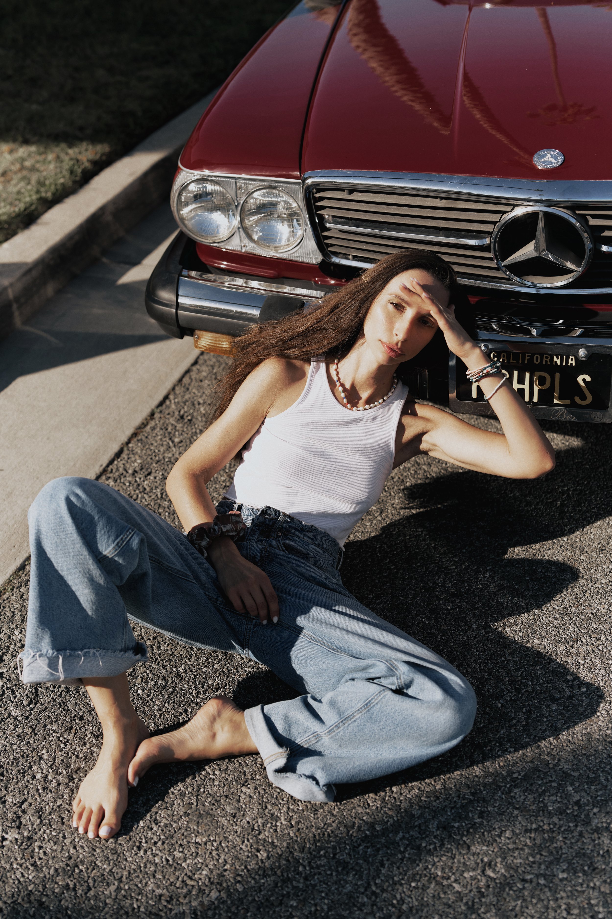 A young woman sits on the pavement next to a parked red Mercedes-Benz car, squinting into the sunlight with her hand on her forehead, in a casual outdoor setting.