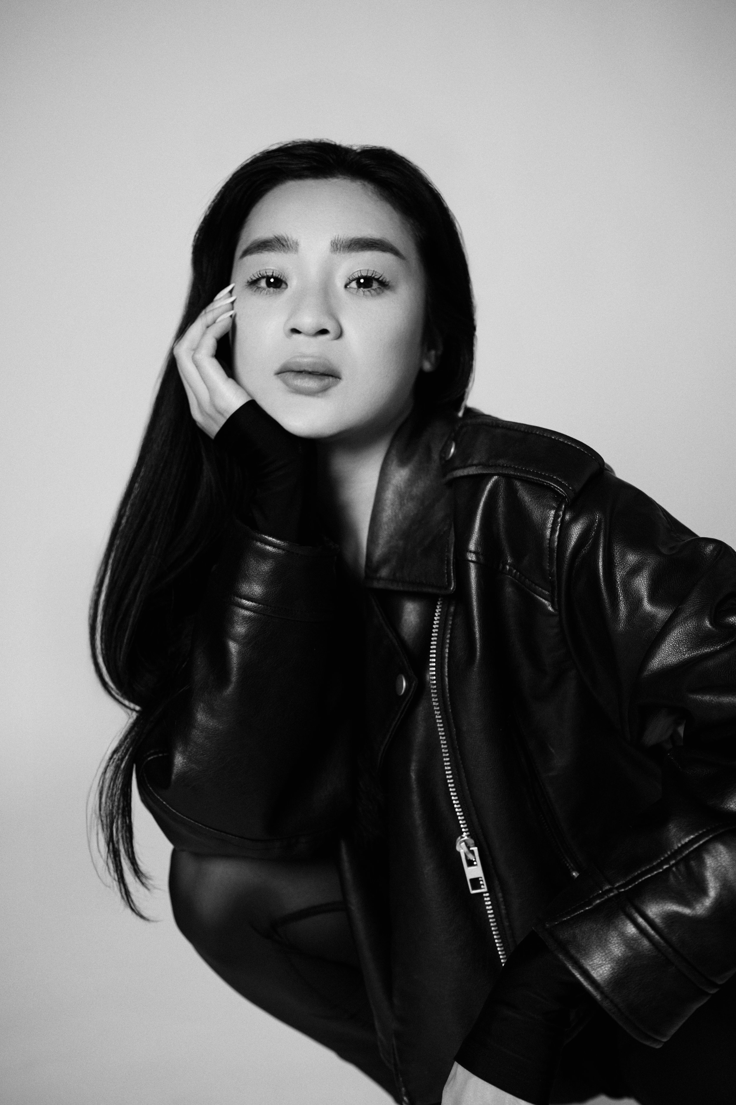 A young Asian woman with long dark hair wearing a black leather jacket, posing with her hand on her face against a plain background.