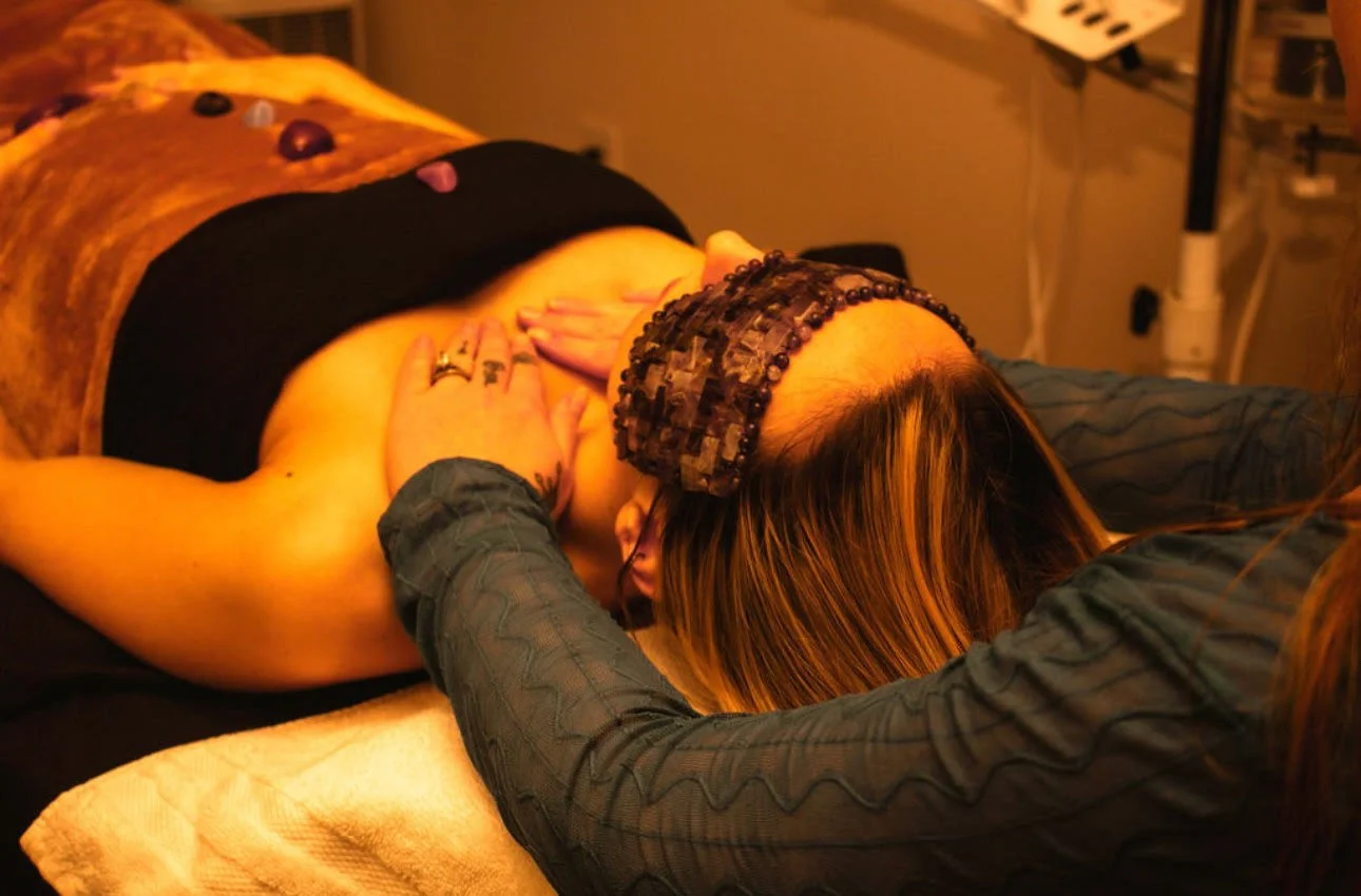 A person receiving a massage, lying face down on a massage table with a dark band around their waist and stones placed on their back, in a dimly lit room.