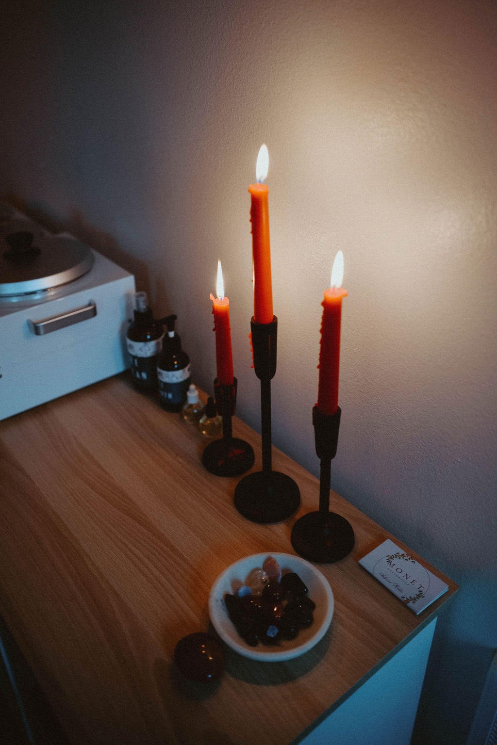 Three candle holders with lit orange candles on a wooden table, next to small bottles and a bowl of assorted chocolates.