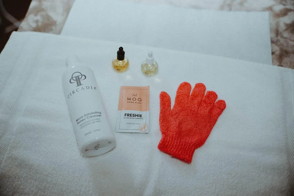 Hygiene and skincare items on a white surface, including a white bottle of Circada delicate exfoliating honey cleanser, two small bottles of oil, a packet of WoW Freshie deodorant, and a red knitted glove.
