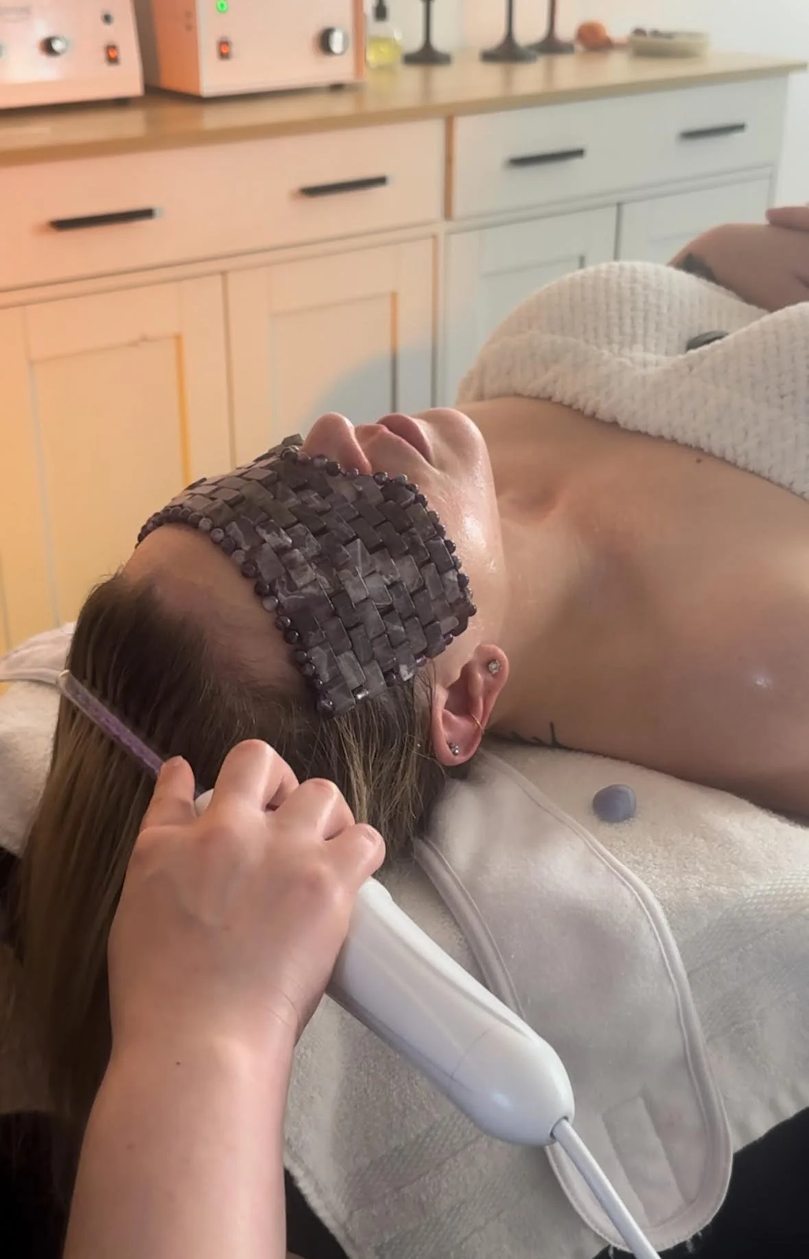 Person receiving a facial treatment with a heated jade roller on their neck, lying down with their eyes closed and a headband, in a spa or therapy room.