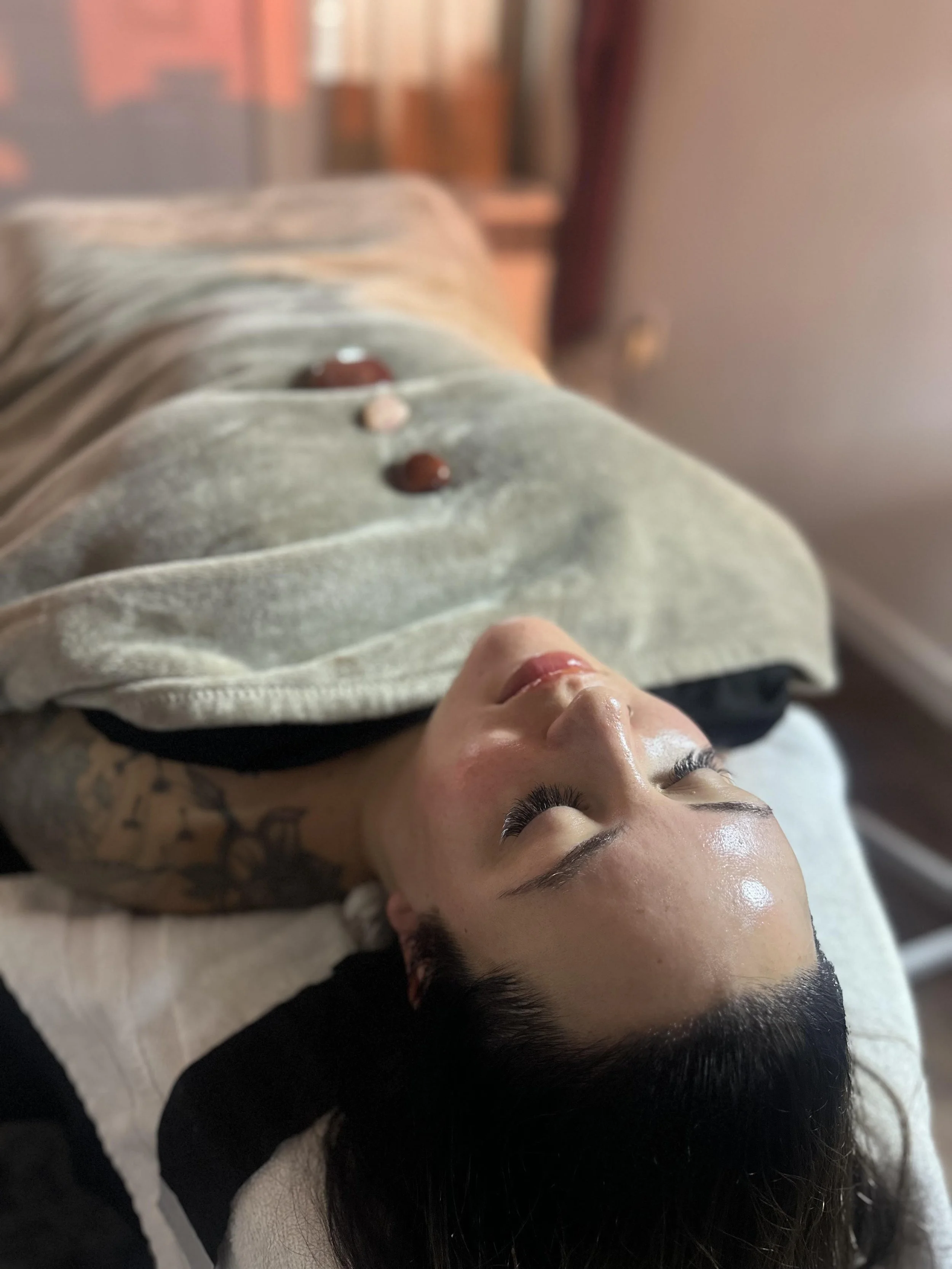 A woman with long dark hair, lying on her back with eyes closed, getting a facial treatment on her face, wearing a black shirt, with a beige towel or blanket covering her body, on a treatment table in a spa or salon.
