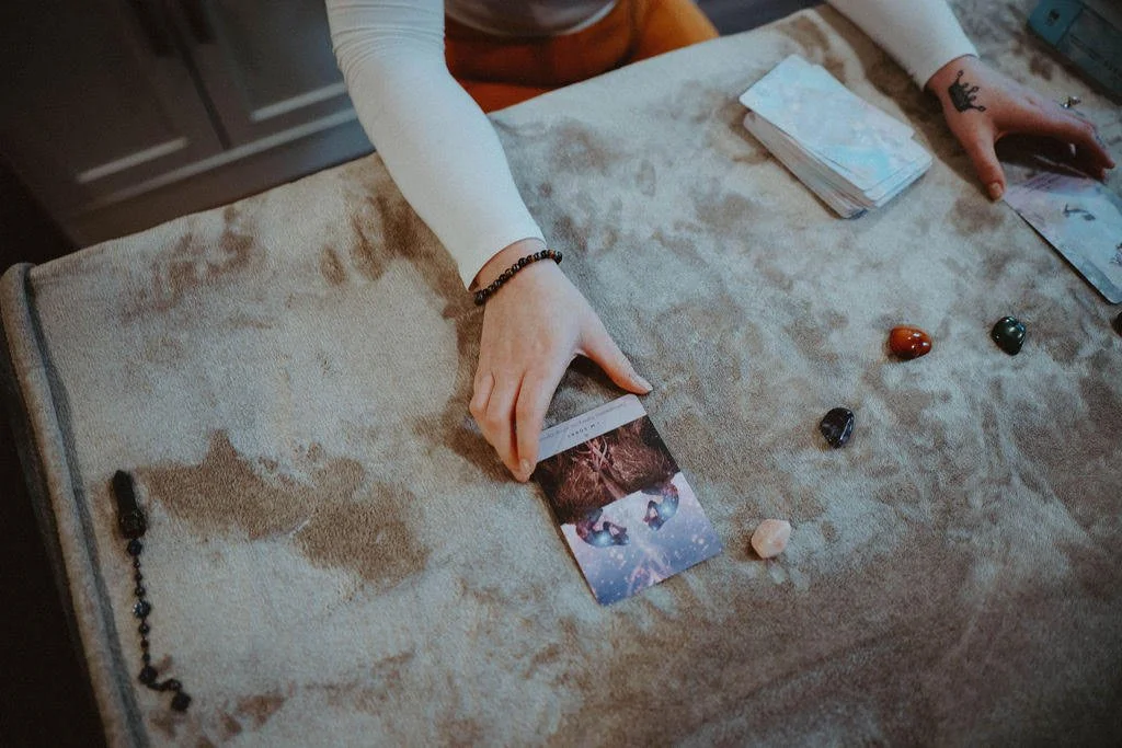 A person wearing a white long sleeve shirt and a black beaded bracelet is sitting at a table with a tarot card spread, deck of cards, and colored stones or crystals on a beige velvet cloth.