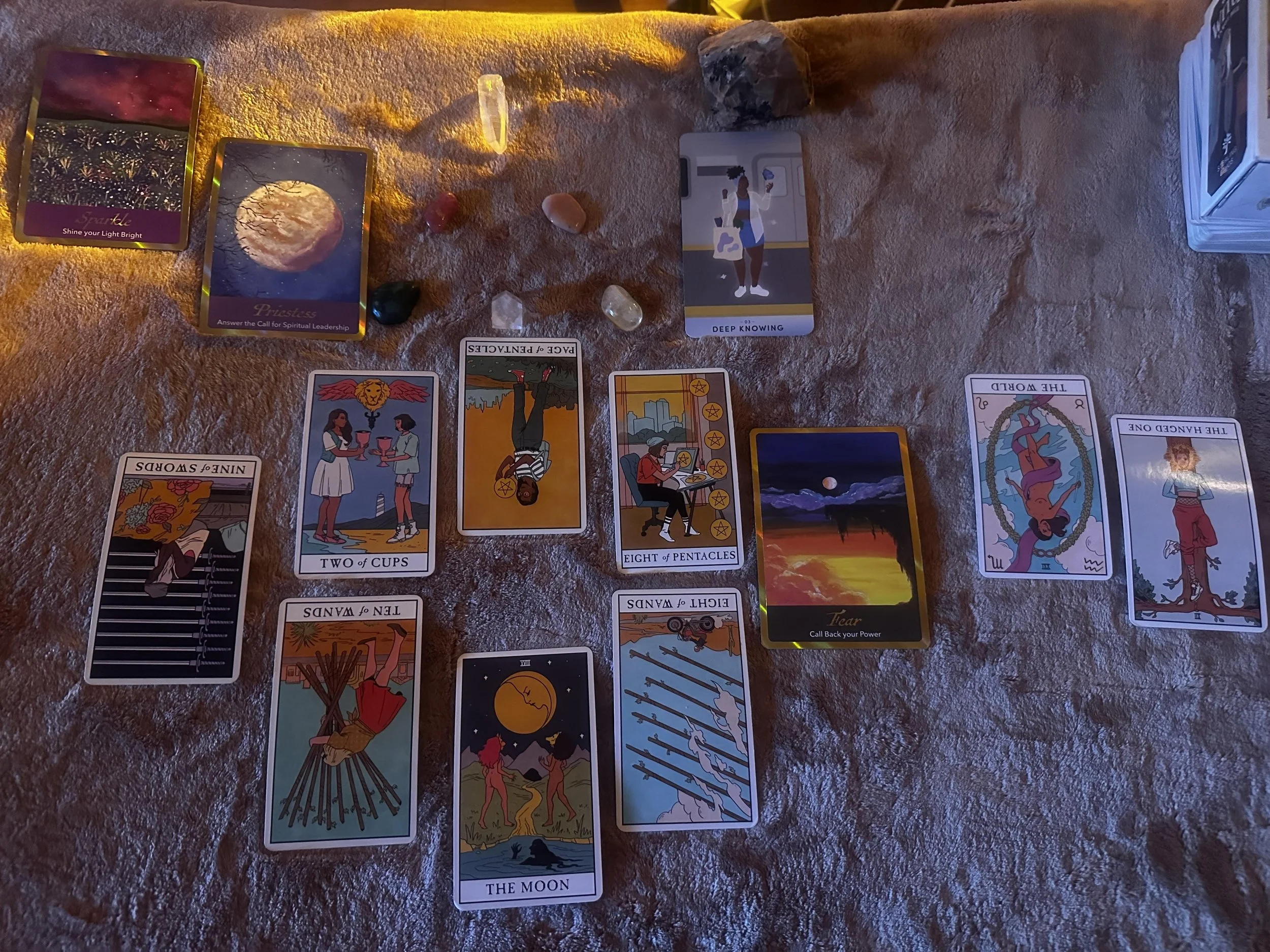 A tarot card reading setup on a textured surface with various tarot cards, crystals, and decorative stones, including the moon, the fool, fear, deep knowing, and other cards arranged in a spread.