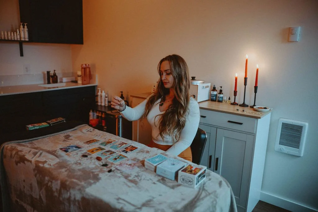 A woman sitting at a table playing a tarot card reading game with tarot cards and game pieces on the table, in a room with candles and various bottles on a cabinet behind her.