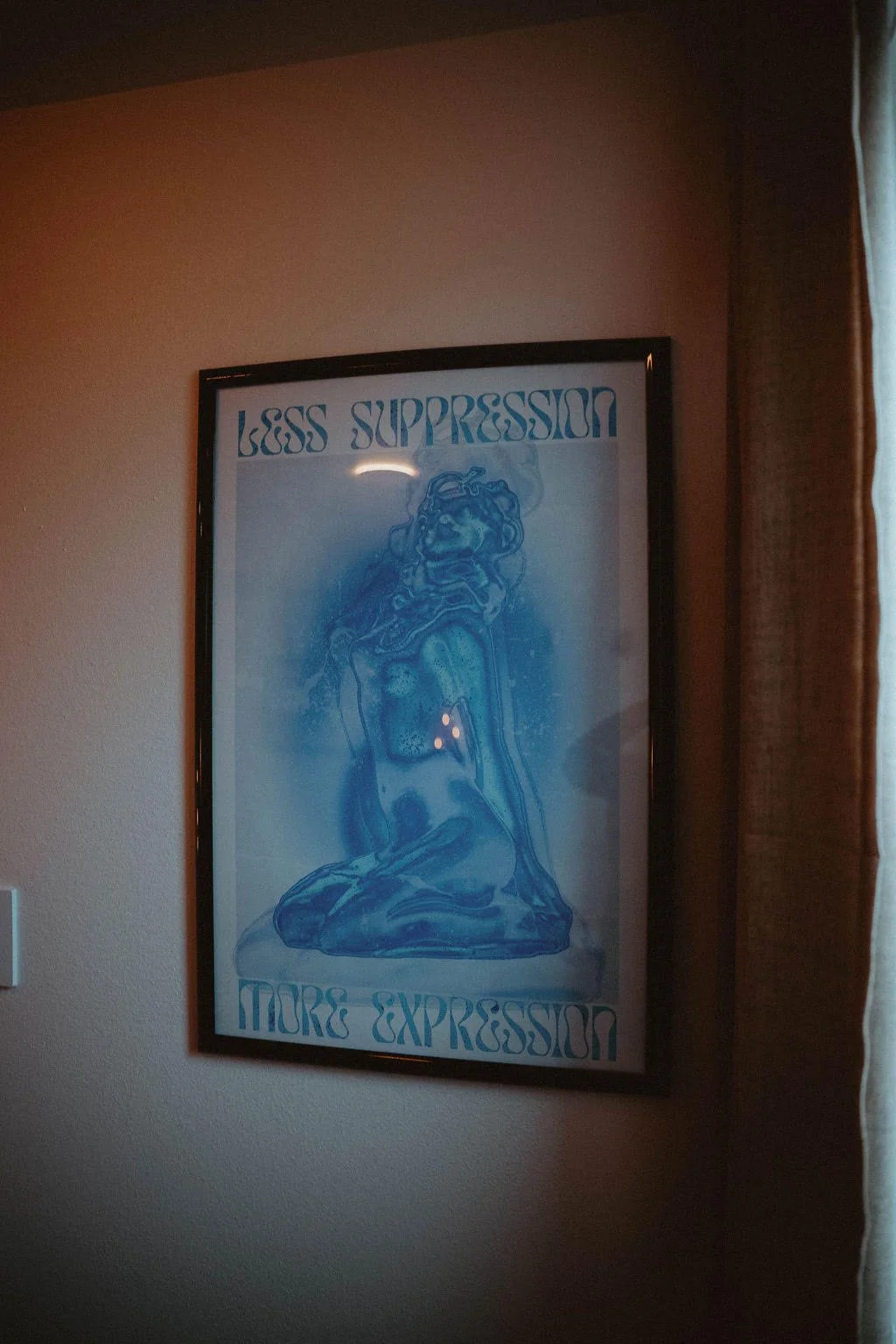 Framed poster on a wall with the text "Less Suppression More Expression" and a blue line drawing of a kneeling female figure.