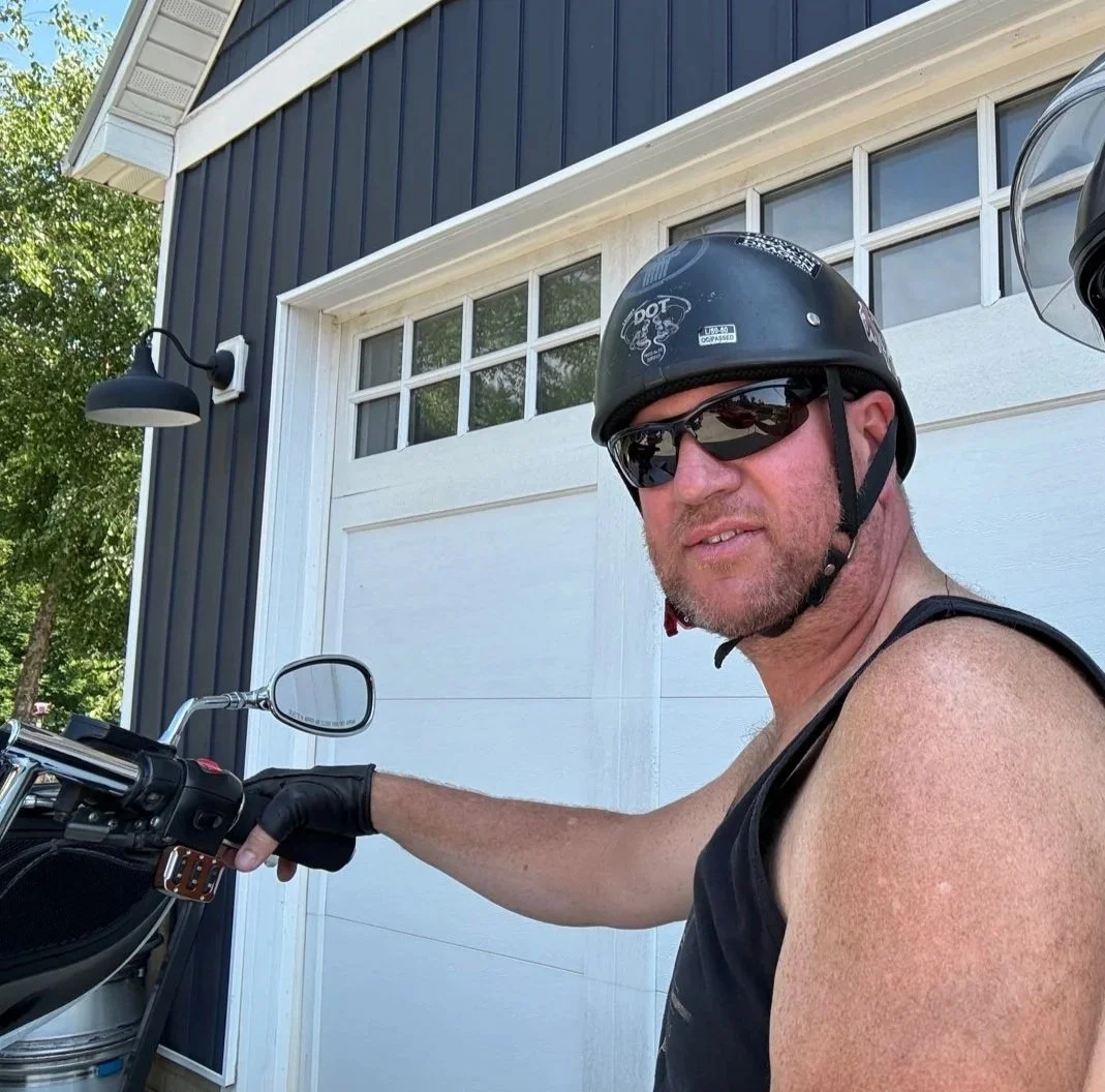A man wearing sunglasses, a helmet, gloves, and a black sleeveless shirt, sitting on a motorcycle in front of a white garage door and a house with blue siding.