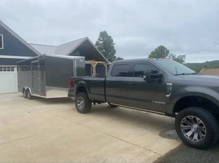 Black pickup truck parked in driveway attached to a large enclosed trailer with a house in the background.