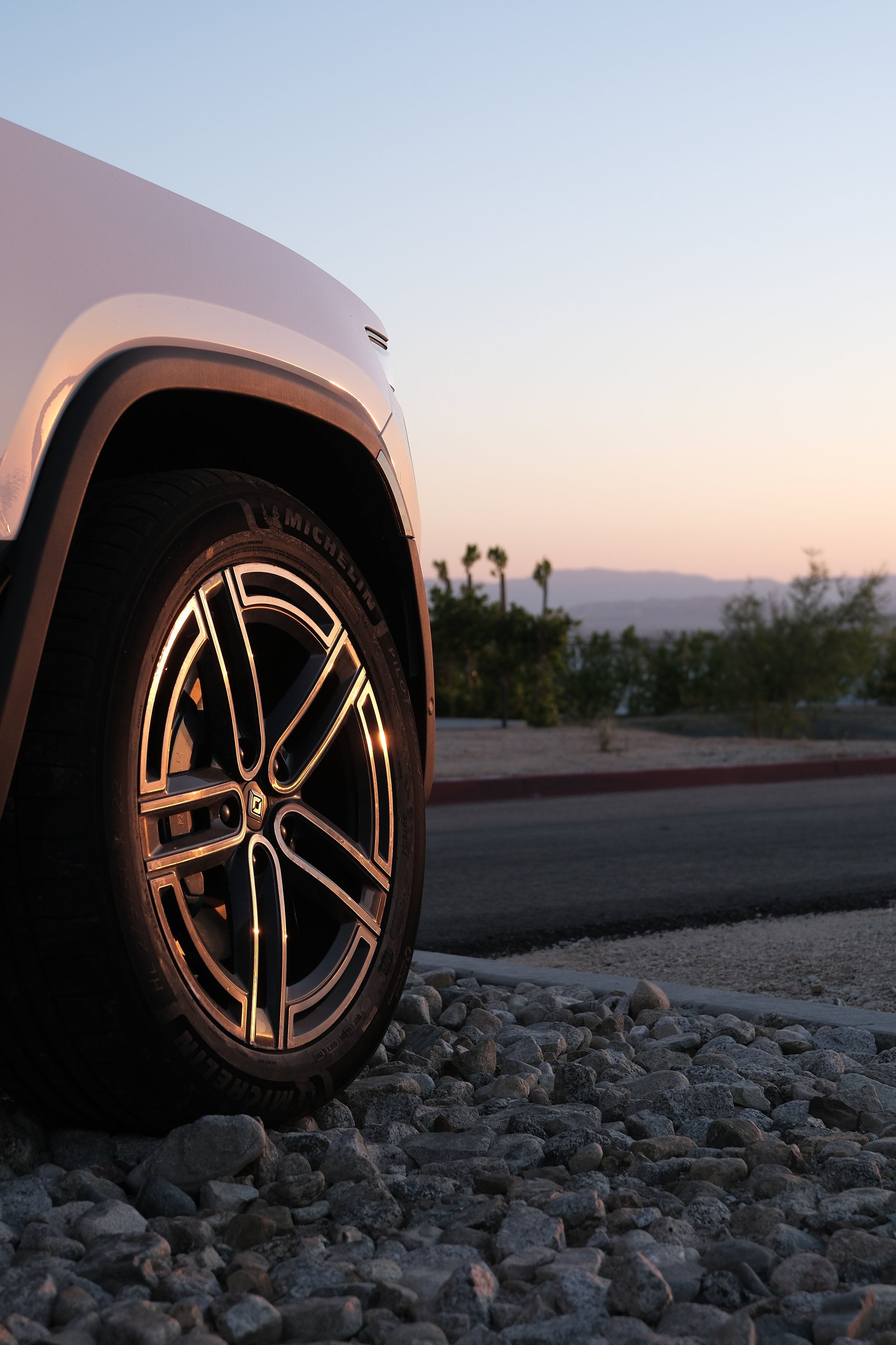 Close-up of a car tire and rim parked on a gravel surface during sunset, with a desert landscape and palm trees in the background.
