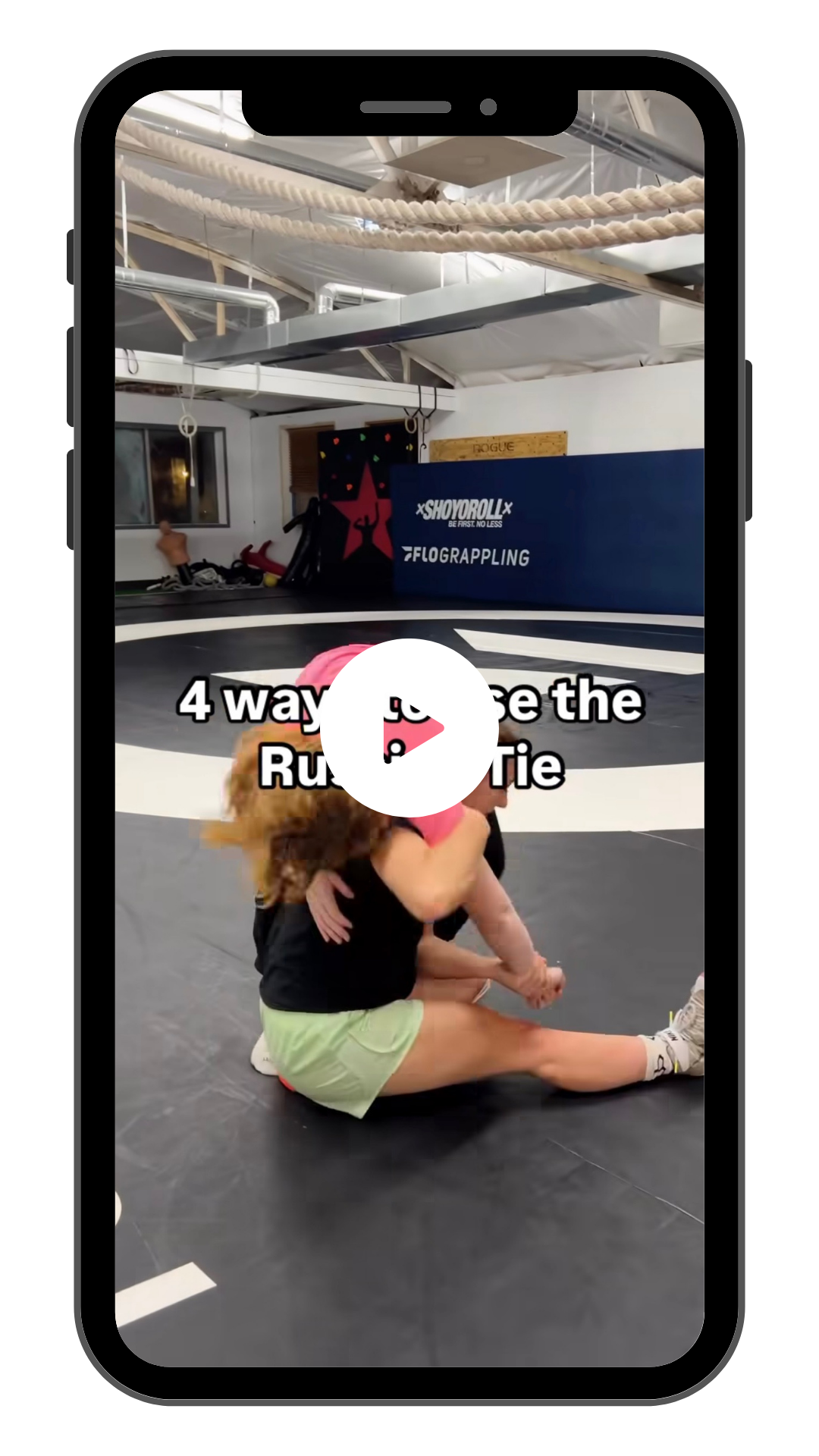 Viral Client Video: Two women wrestling on a mat in a gym with gym equipment and posters in the background. Text overlay reads "4 ways to use the Russian Tie." and a play button icon on a mobile device screen.