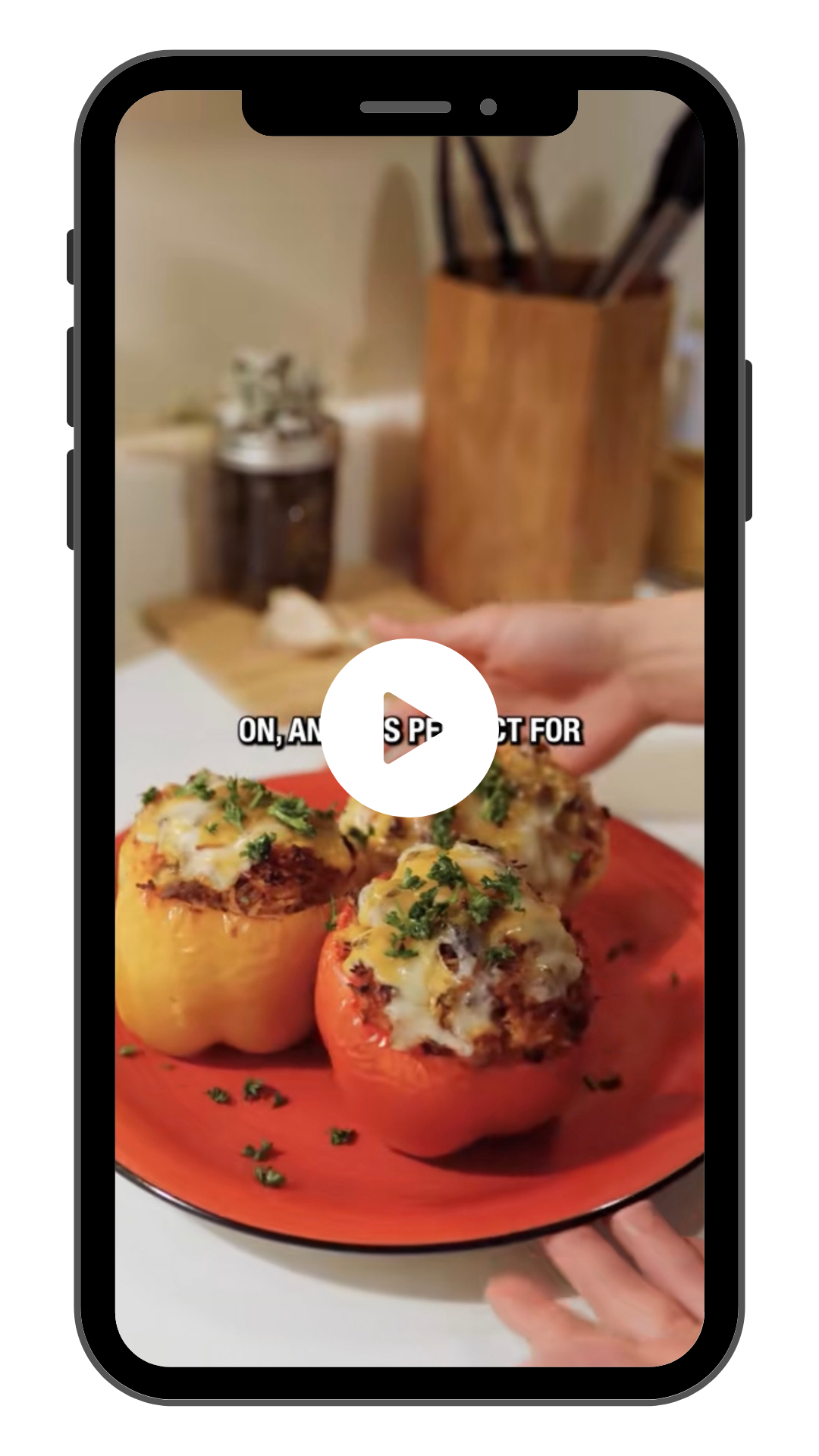 Client Video: Close-up of stuffed peppers topped with cheese and herbs on a red plate, with a blurred kitchen background and a play button icon on a mobile device screen.