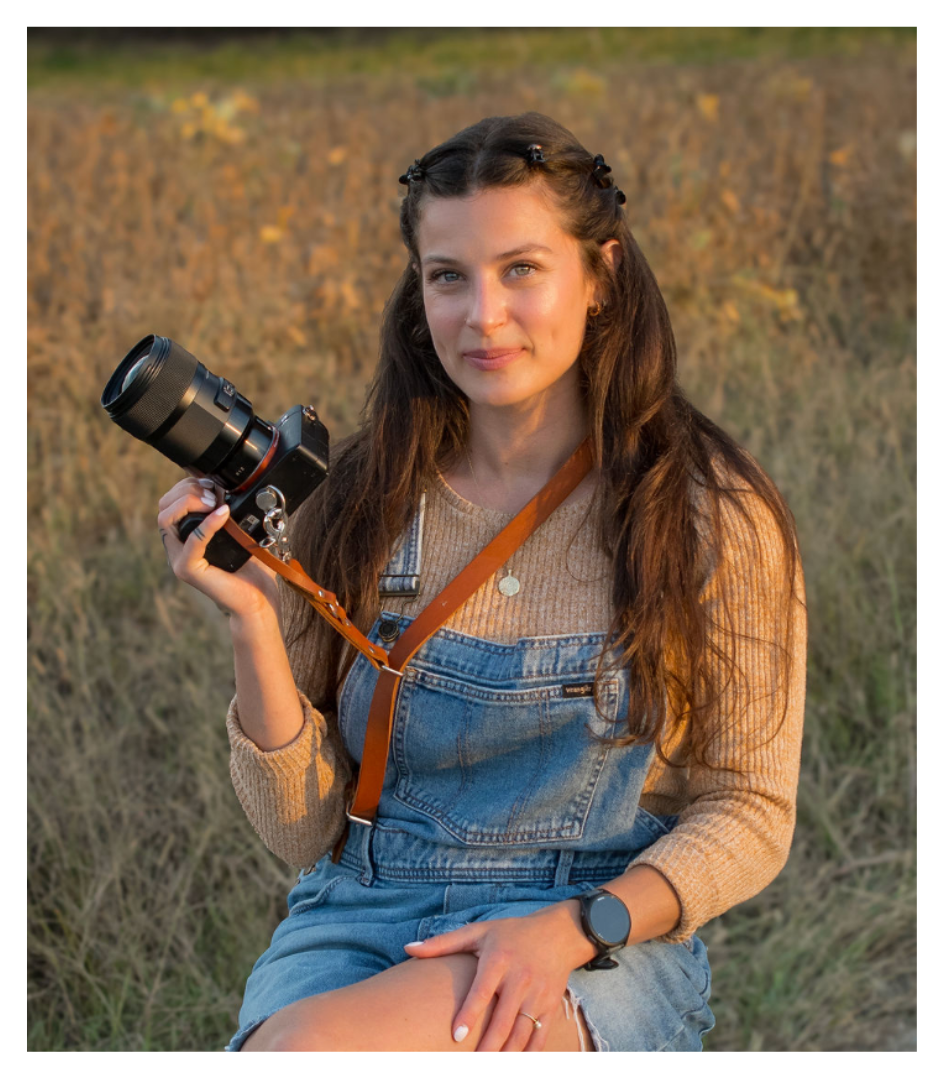 Young woman sitting outdoors in a field, holding a camera, wearing denim overalls, a beige sweater, and a smartwatch, during golden hour.