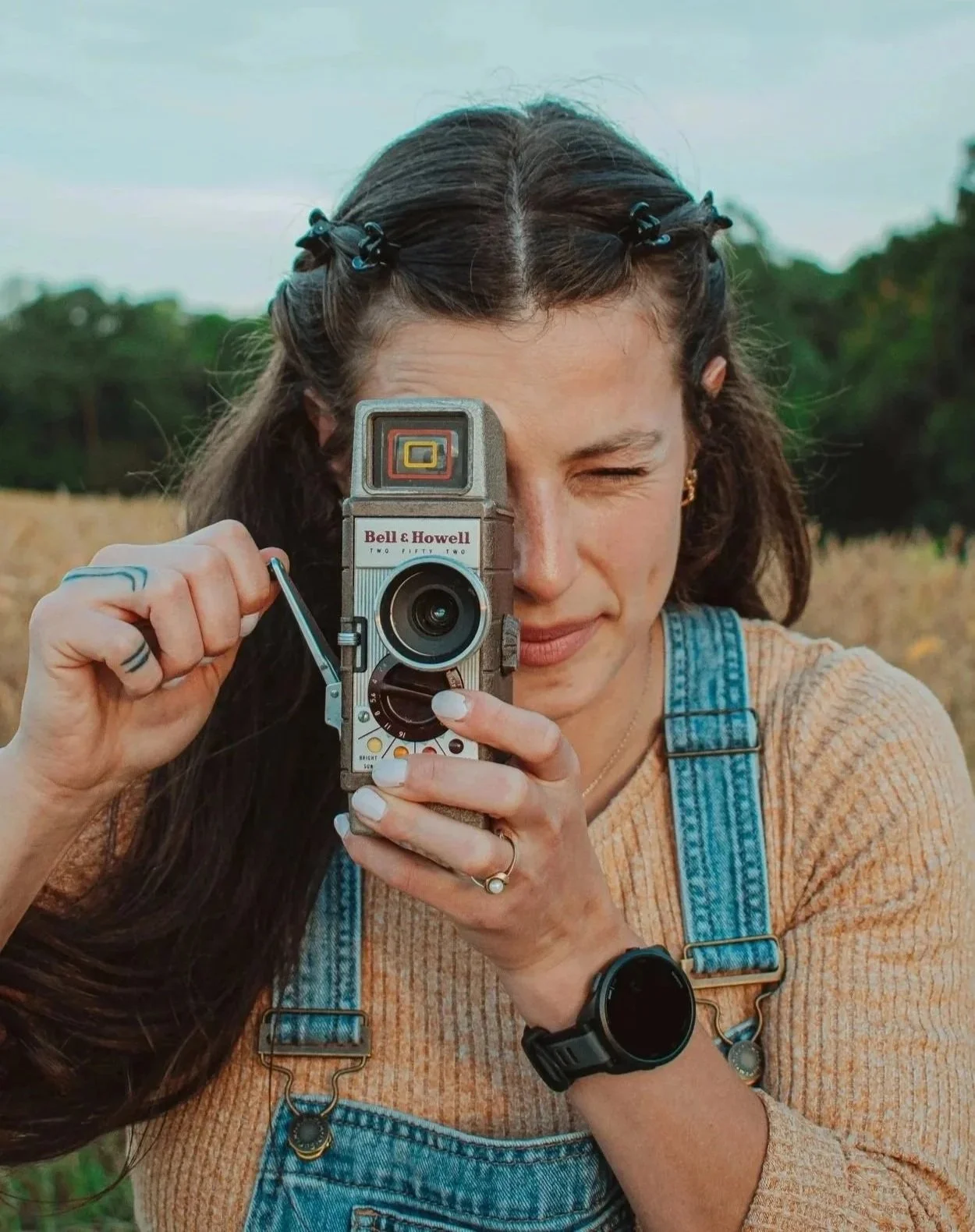 A woman with long dark hair, wearing a beige sweater and denim overalls, takes a photo with a vintage camera in an outdoor setting with trees and grass in the background.