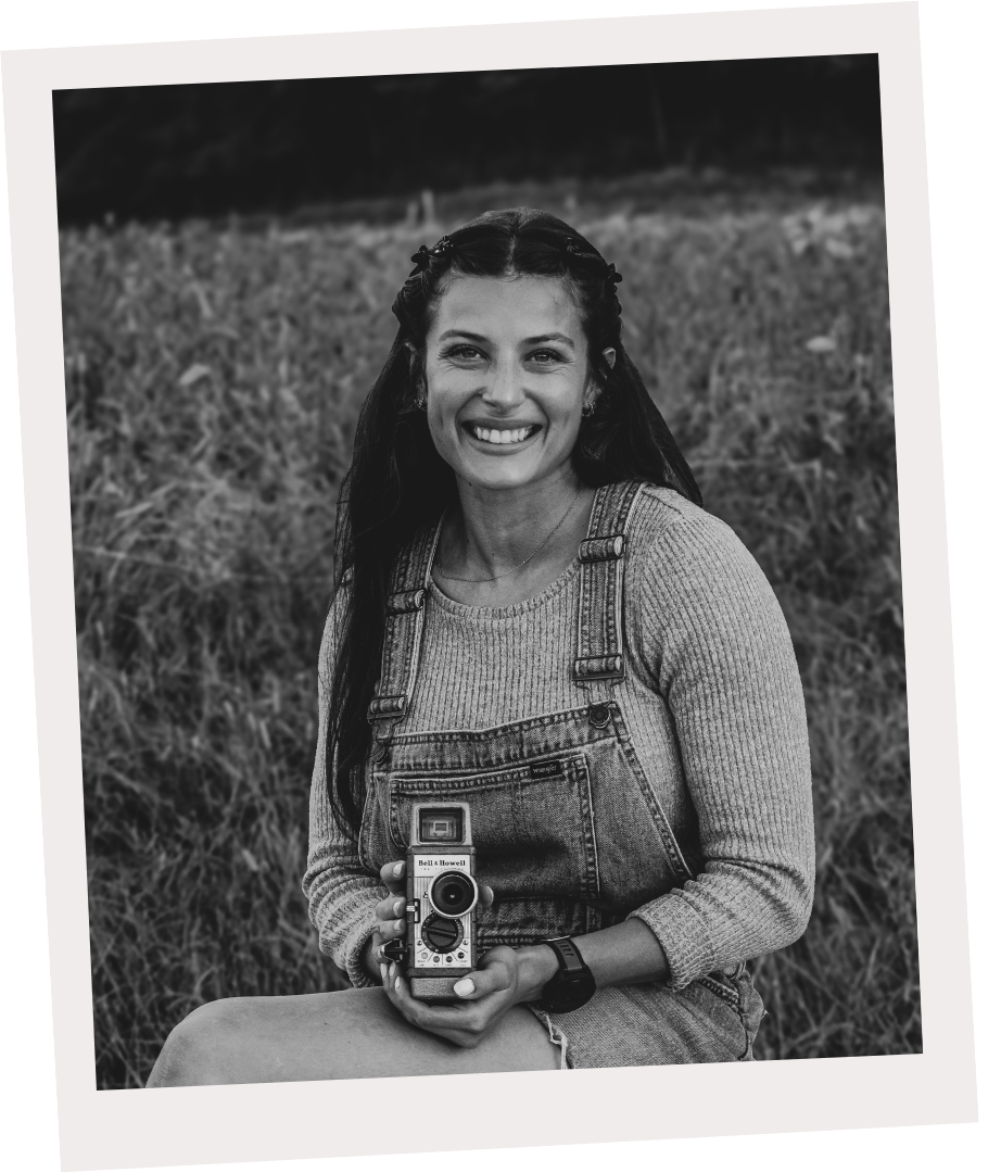 A smiling woman sitting outdoors on grass, holding a vintage camera, wearing a long sleeve top and overalls.