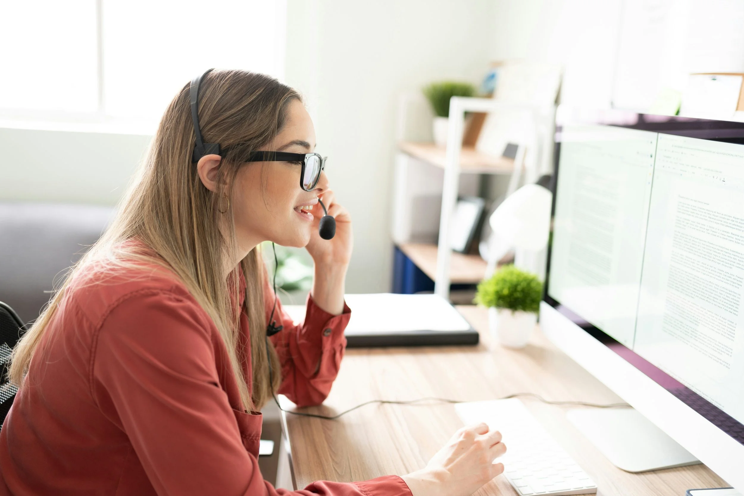 Woman smiling in a red shirt working at a computer with a headset in a bright modern office.