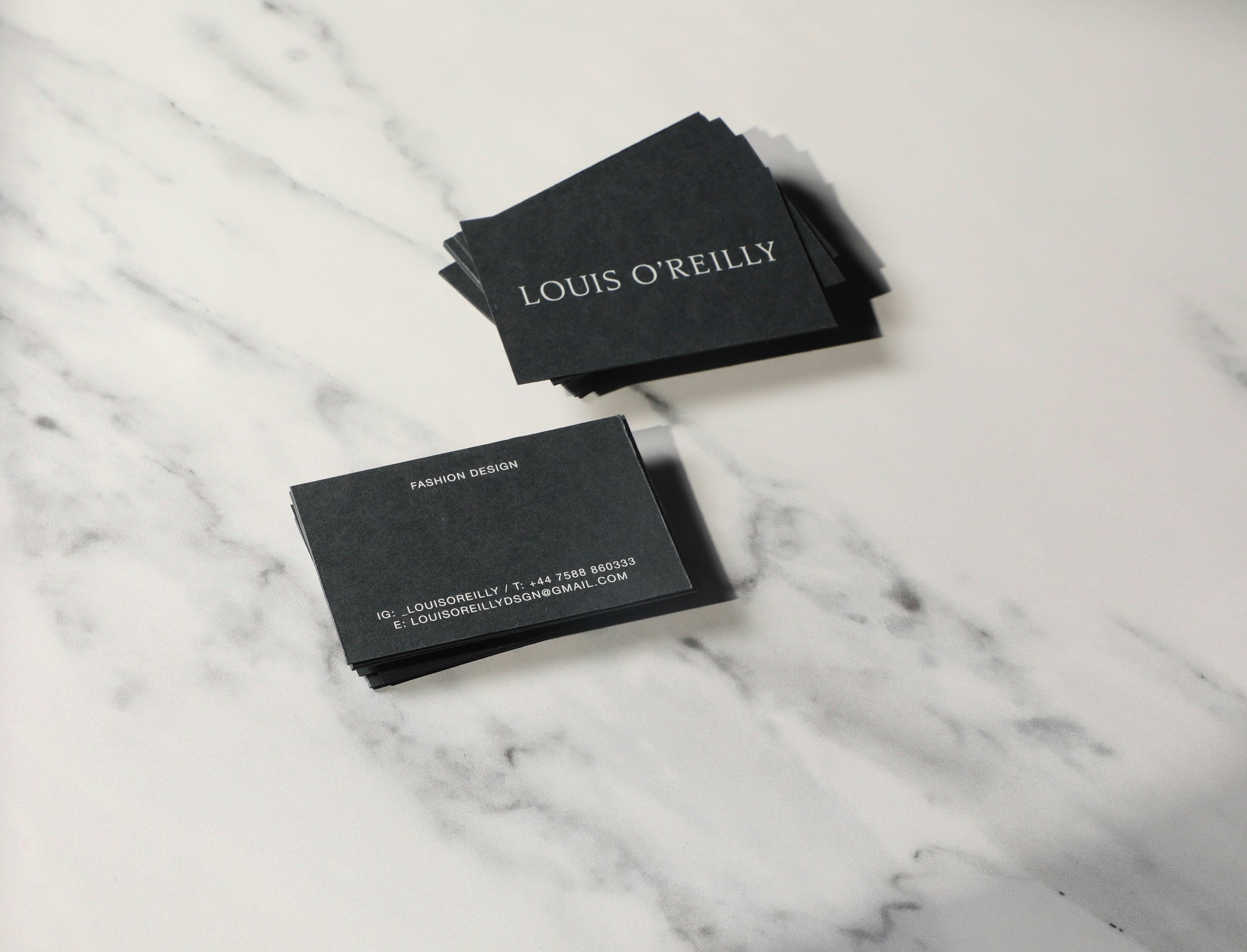 Stack of black business cards with white text, placed on a white marble surface. One card shows the name 'LOUIS O'REILLY' and another indicates 'Fashion Design' with contact details.