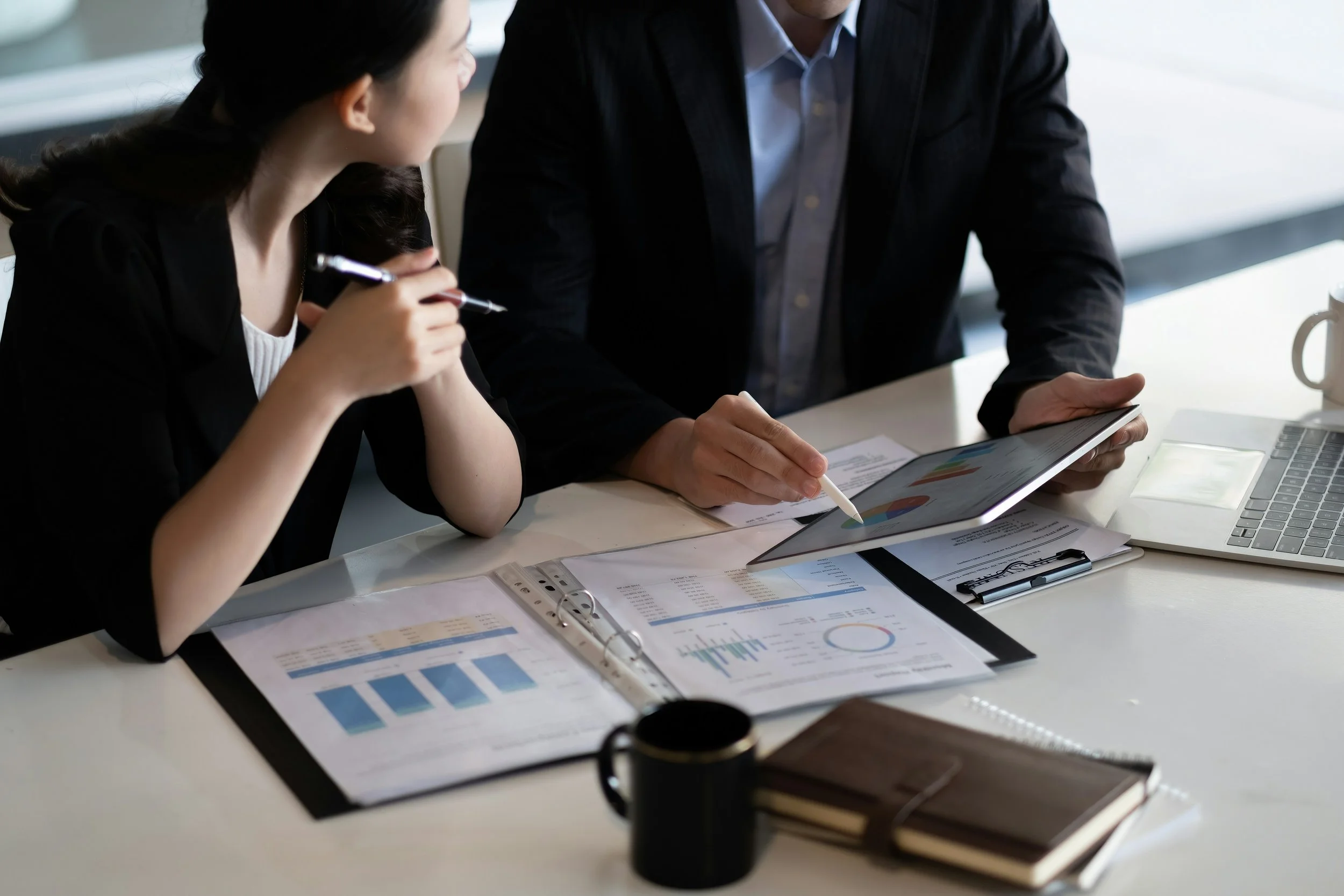 Two people discuss business documents and data charts in a meeting, with a tablet, laptop, and coffee mug on the desk.