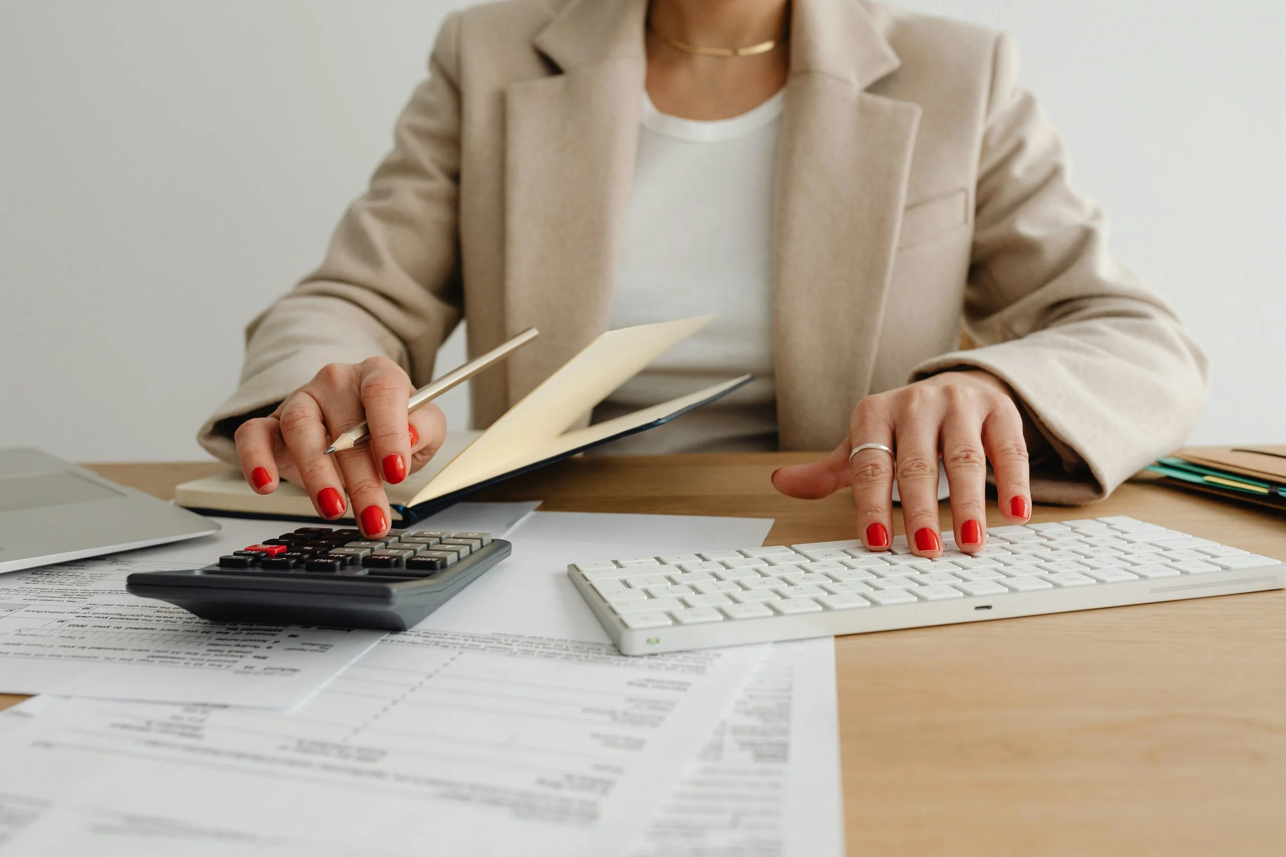 A woman in a beige blazer using a calculator, typing on a white keyboard, writing in a notebook, with documents and a laptop on a desk.