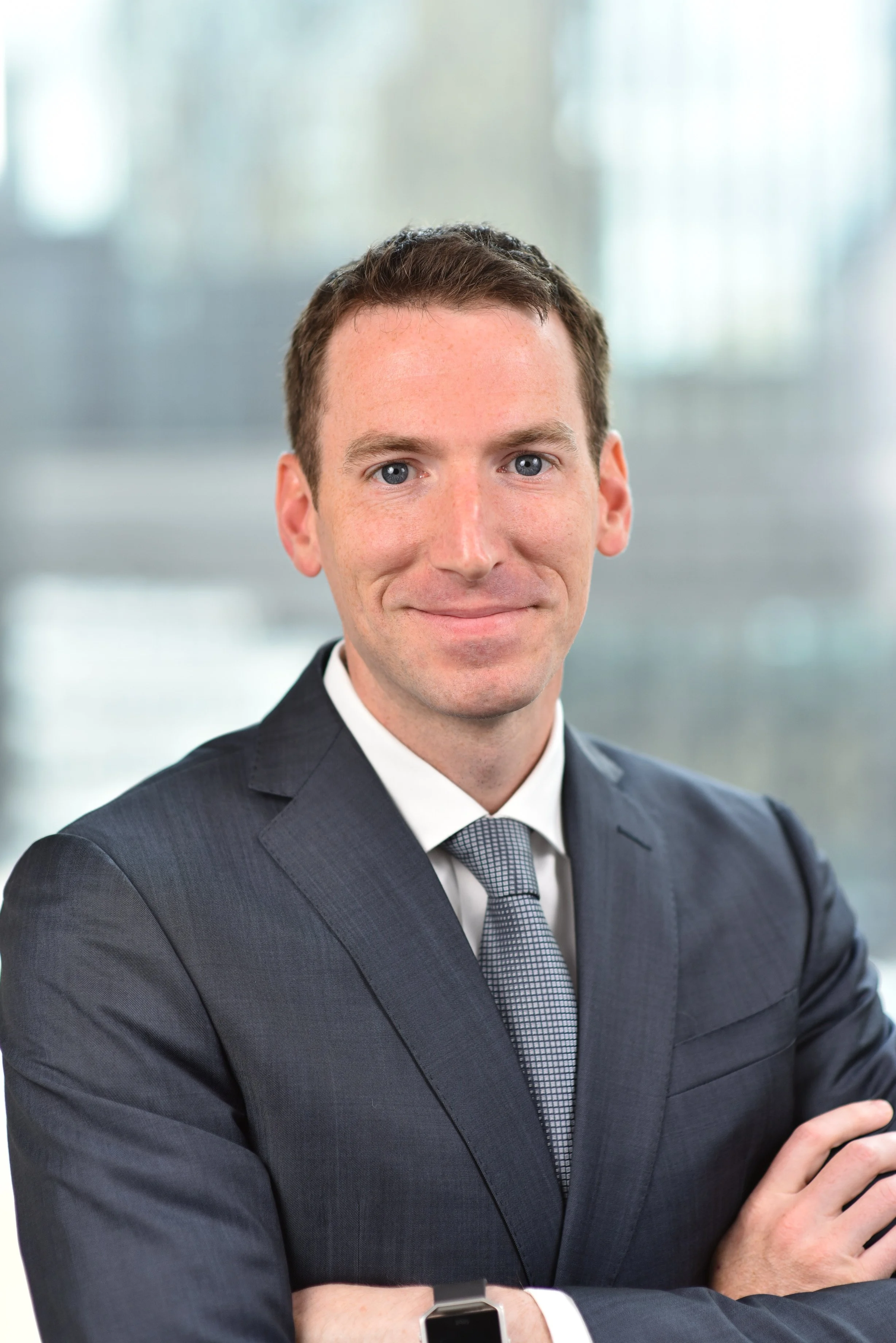 A professional man in a dark gray suit, light tie, and white shirt, standing with his arms crossed, in an office setting with bright, diffused natural light.