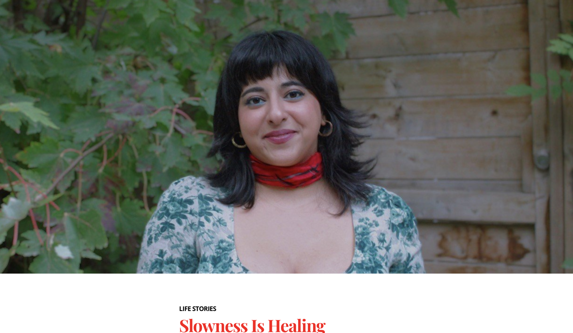 A woman with dark hair, wearing a floral top and red scarf, standing outdoors in front of green foliage and a wooden fence, smiling at the camera.