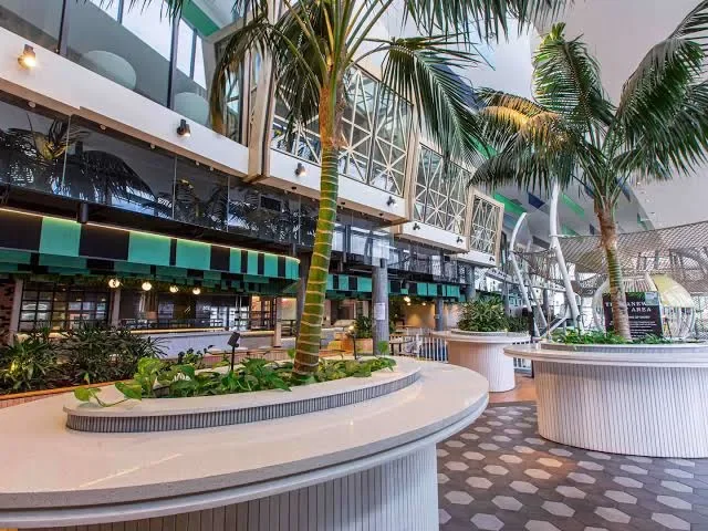 Indoor shopping mall with plants, palm trees, and storefronts, featuring modern architecture with large glass windows and a patterned floor.