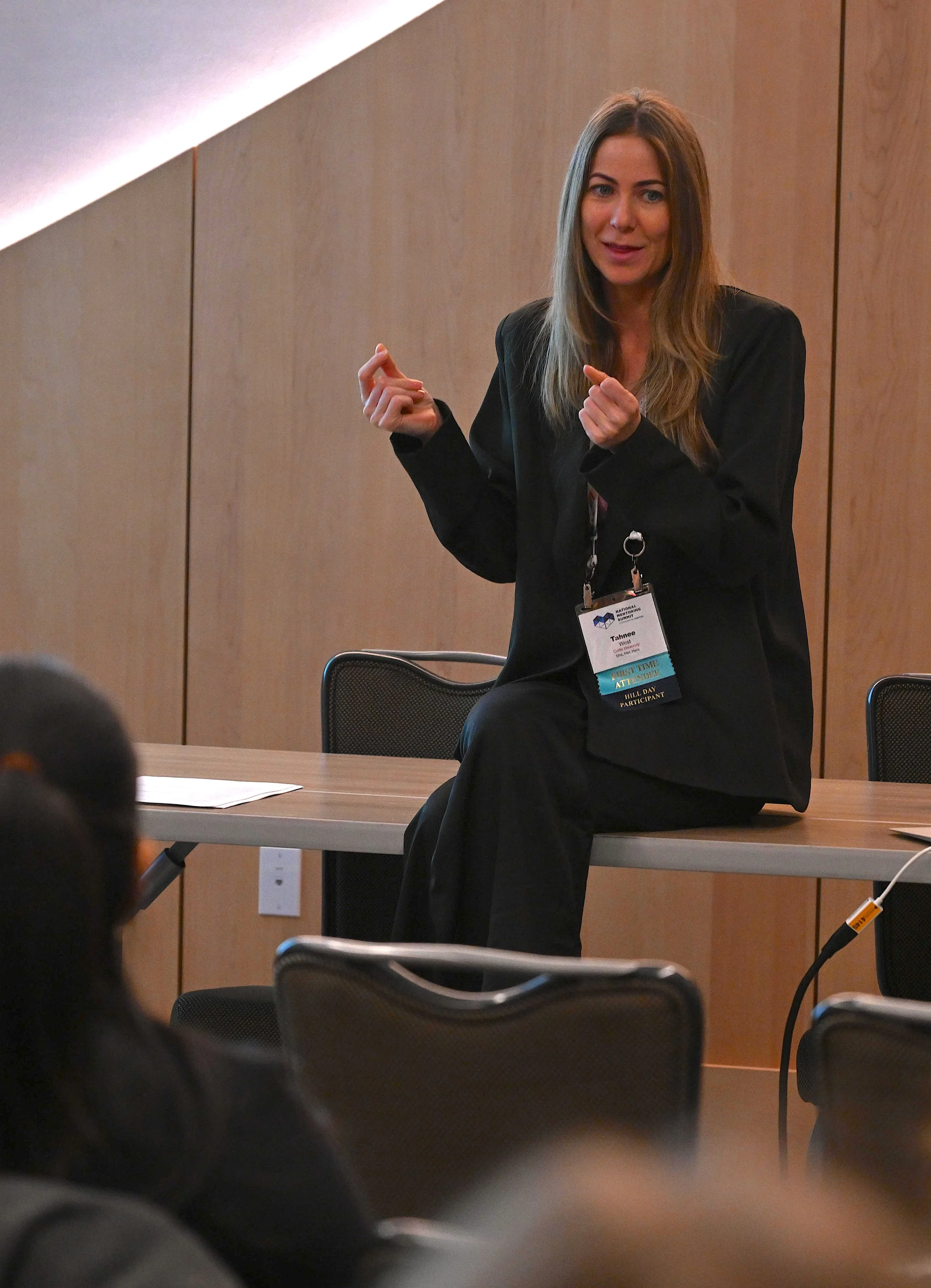 A woman with long brown hair, wearing a black blazer and a name tag, sitting on a gray chair at a conference or seminar, gesturing with her hands while speaking.