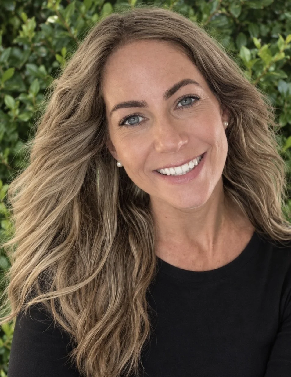 A woman with long wavy light brown hair, blue eyes, and pearl earrings, smiling outdoors with green shrubbery in the background.